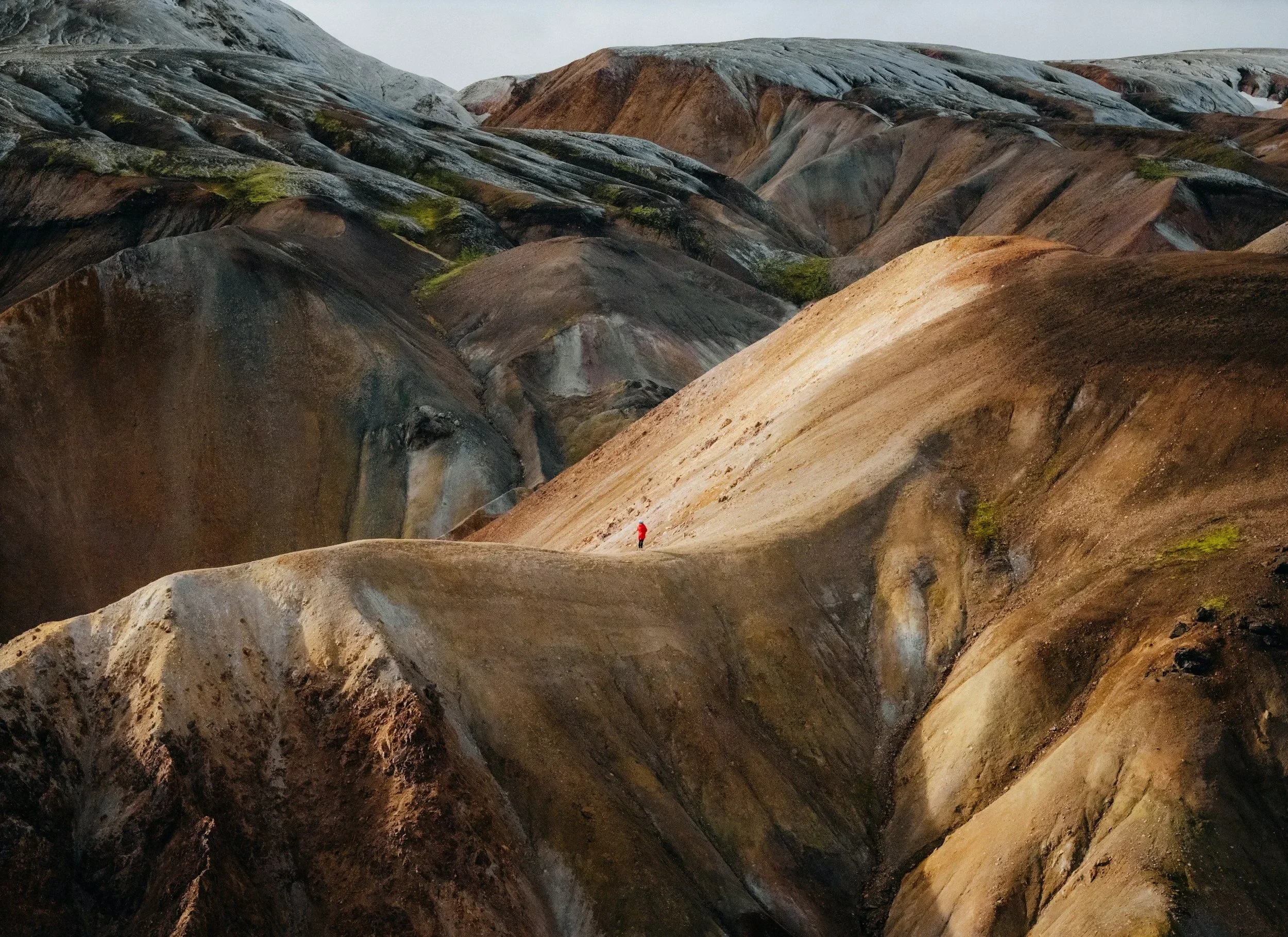 A lone hiker in a bright red jacket walks across colorful volcanic hills, showcasing dramatic earth tones and rugged terrain in a remote, otherworldly landscape—perfect for travel, adventure, and nature-themed content.