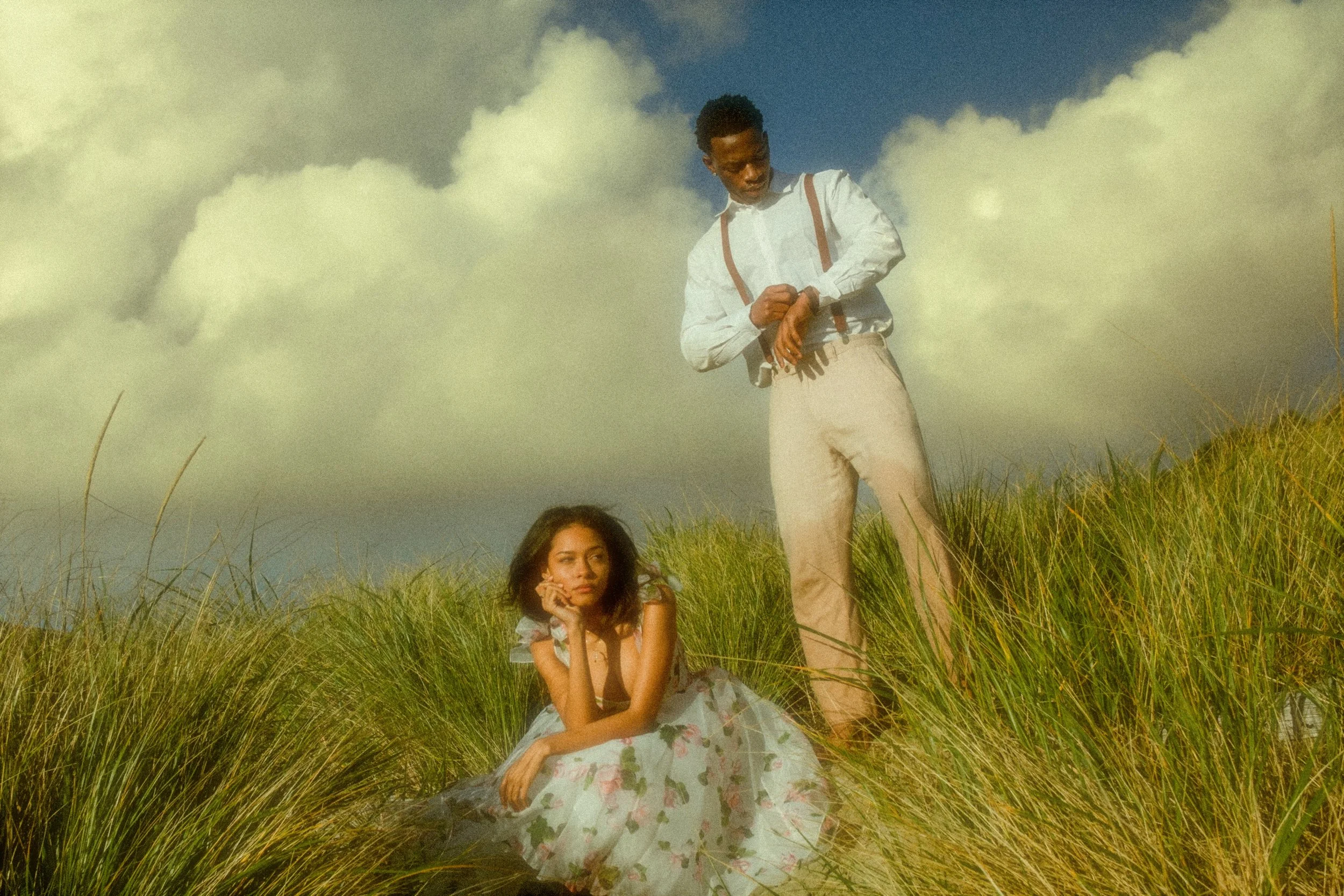 A young man standing in a grassy field with a girl sitting on the ground in front of him, both under a partly cloudy sky.