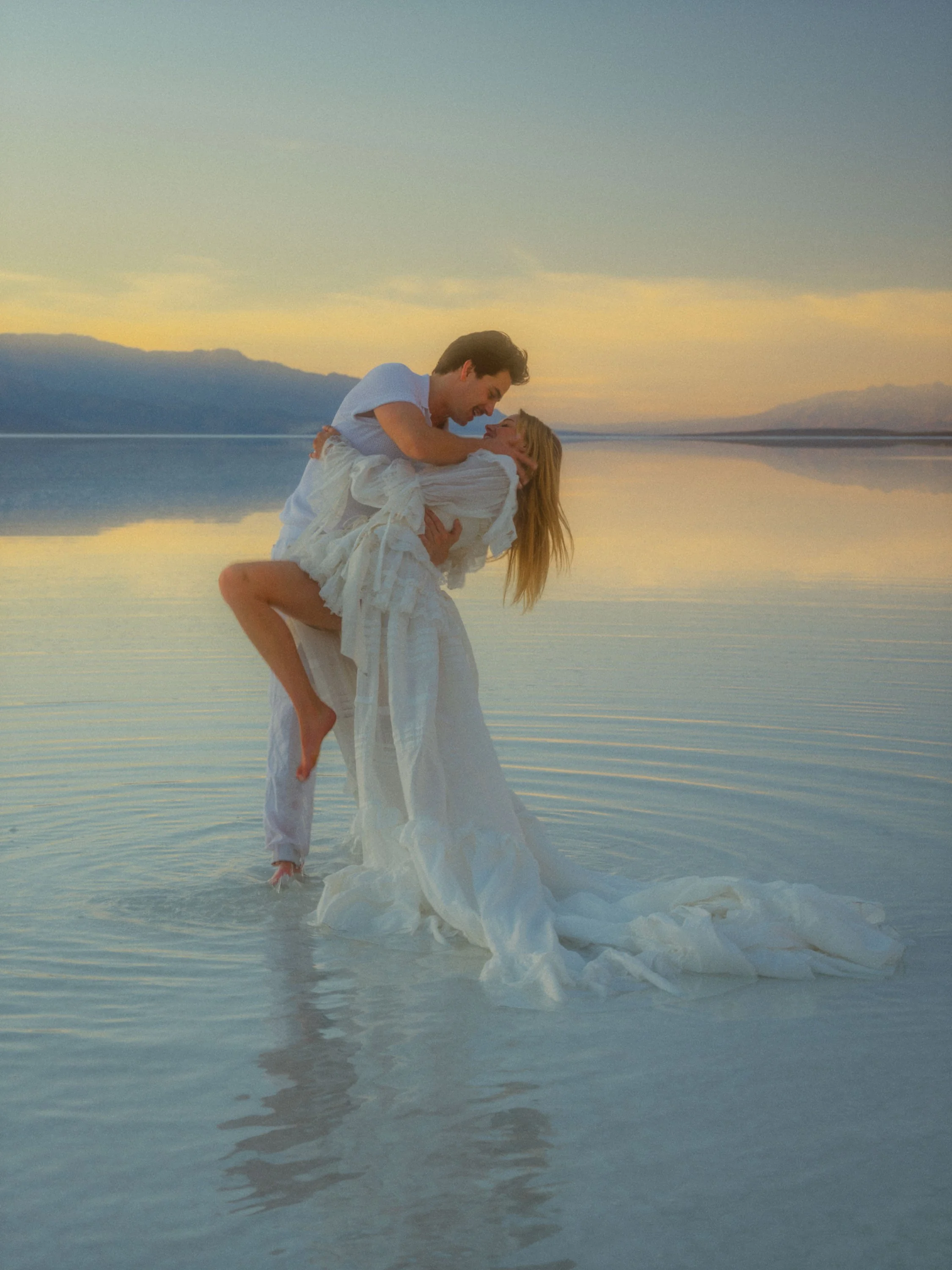 A man lifting a woman in a white dress in shallow water at sunset, with mountains and a cloudy sky in the background.