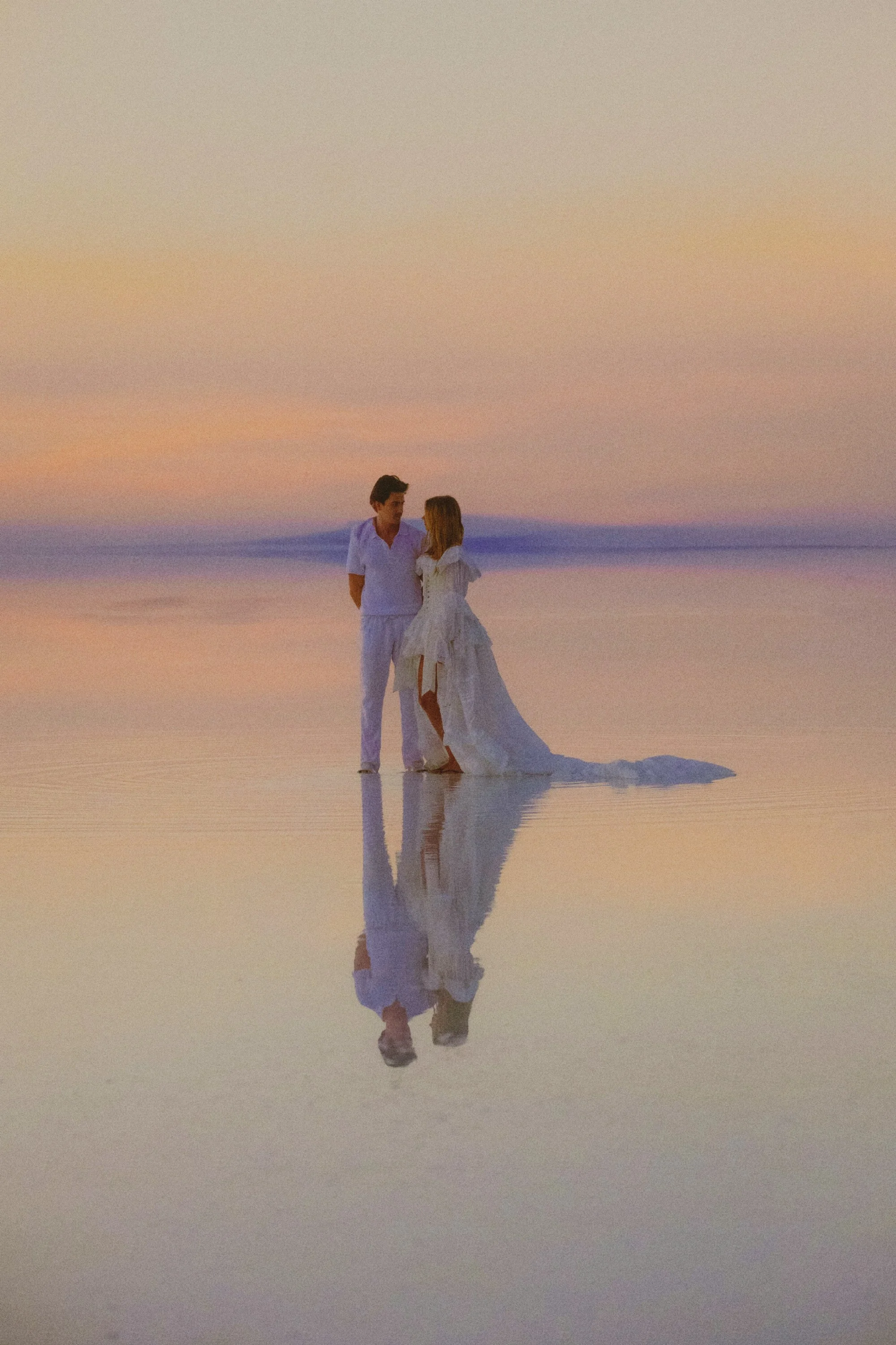 A couple dressed in white clothes, standing close together on a reflective water surface during sunset, with soft pastel colors in the sky.