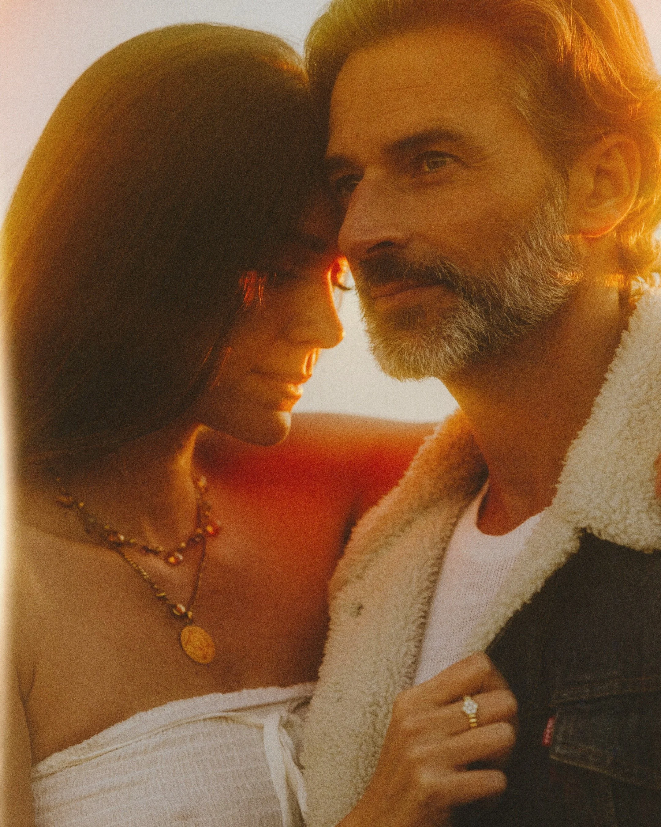 A close-up photo of a couple: a woman with long dark hair, jewelry, and a white sweater, and a man with gray hair and a beard, wearing a fleece jacket, looking into each other's eyes, with warm lighting.