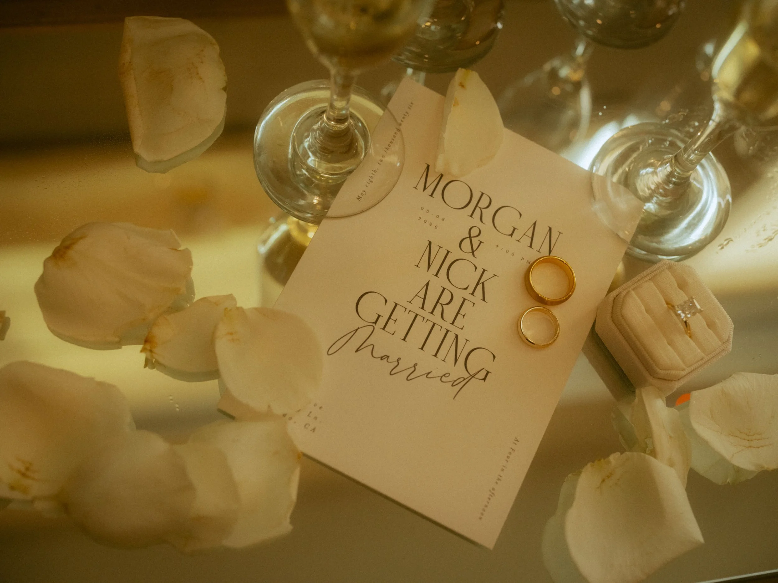 A wedding invitation card with a white background, gold rings, and white shells, surrounded by seashells. The invitation reads 'Morgan & Nick are getting married' and includes the date '05.08.2026'.