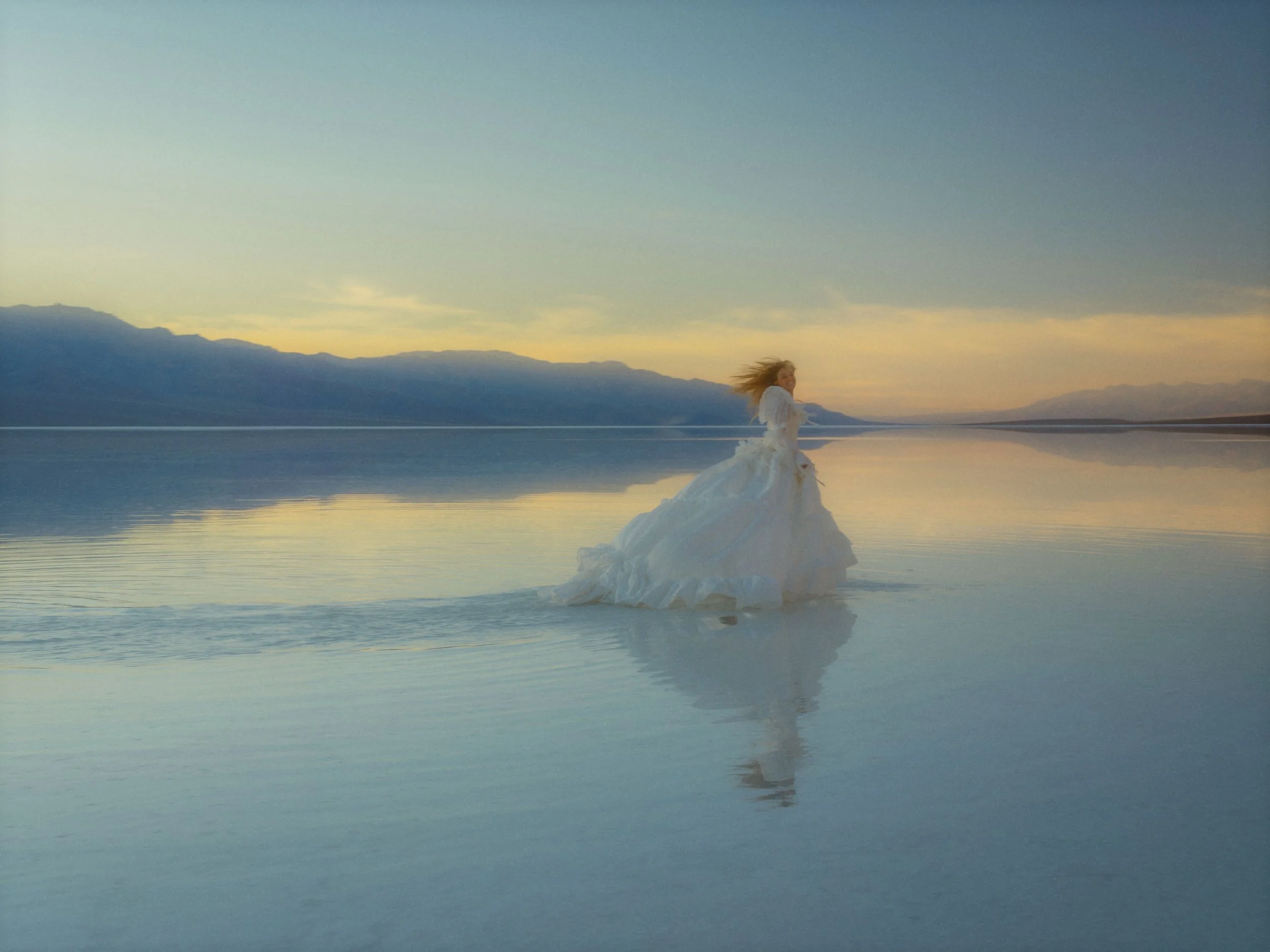 A woman in a white dress stands on a small iceberg or piece of ice floating on a calm body of water during sunset or sunrise, with mountains visible in the background.