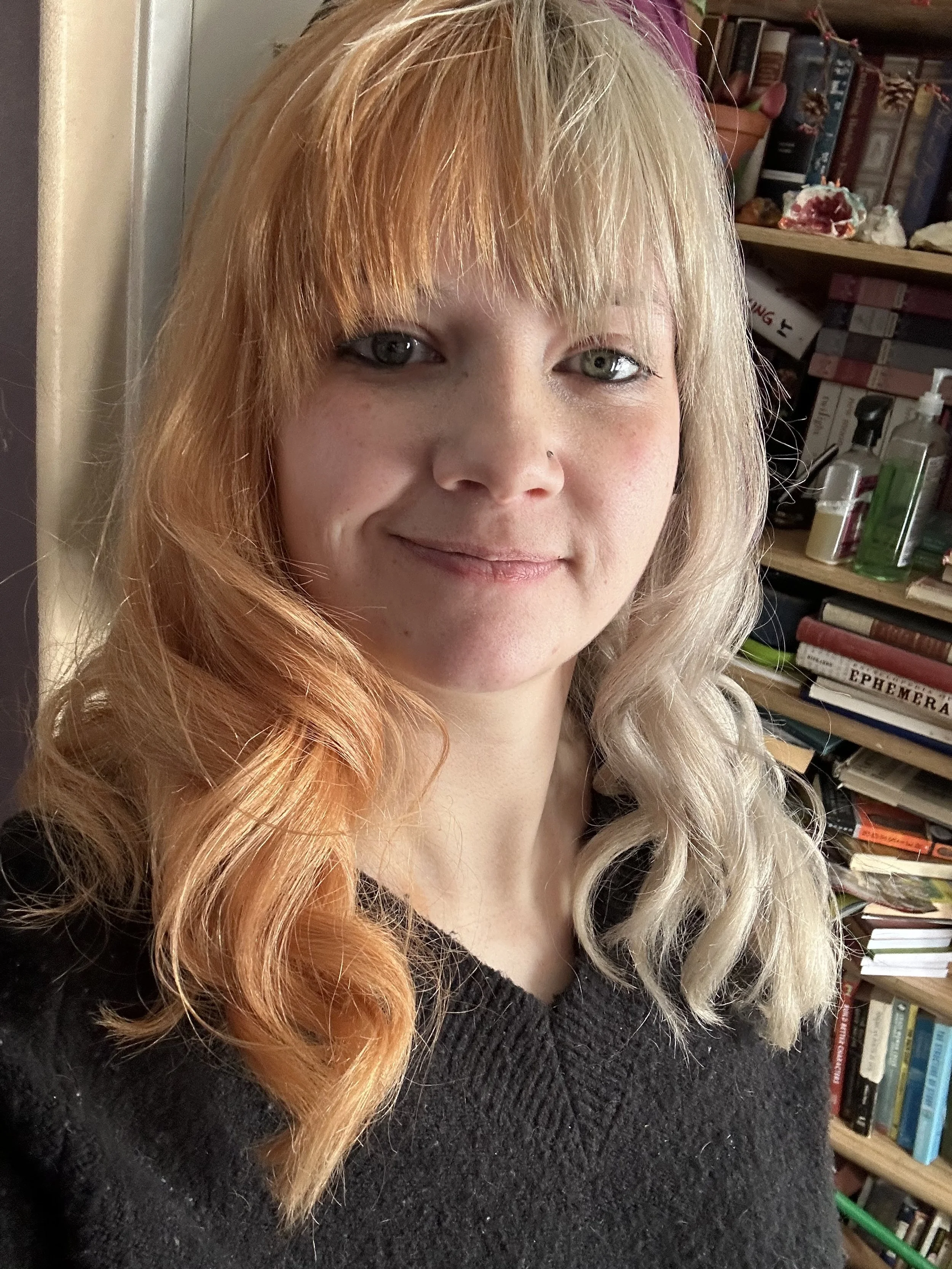 A woman with orange and blonde hair, and green eyes, smiling in front of a bookshelf containing books and decorative items.