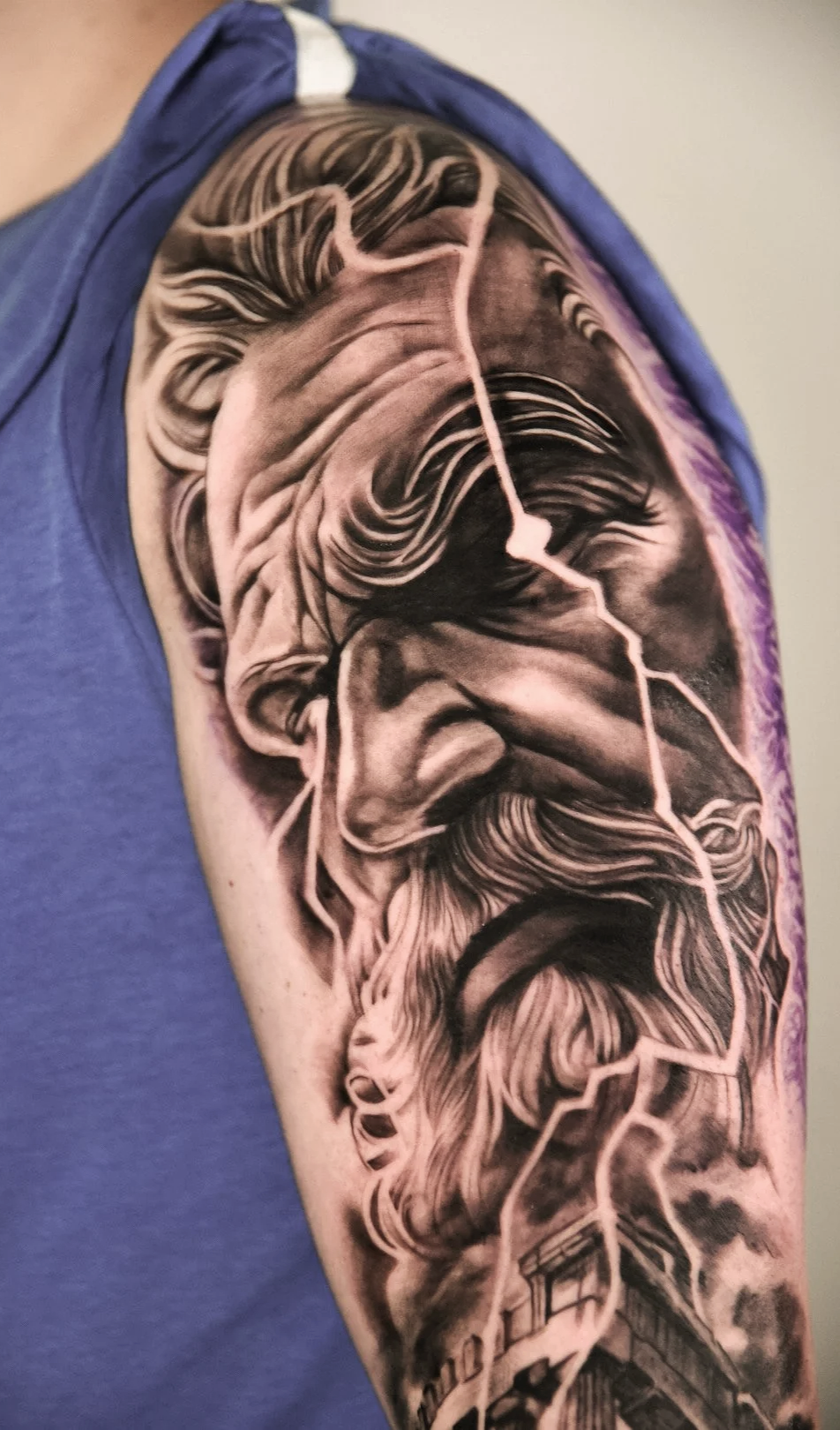 Black and gray tattoo of a bearded man's face with long hair and an orange lightning bolt behind, located on the arm of a person wearing a blue t-shirt.