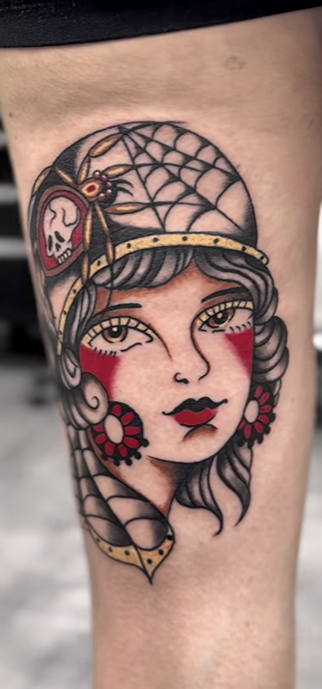 A tattoo of a woman with wavy hair, wearing a gold headband with a spider, a dancer's mask, and large red earrings, in a traditional style.