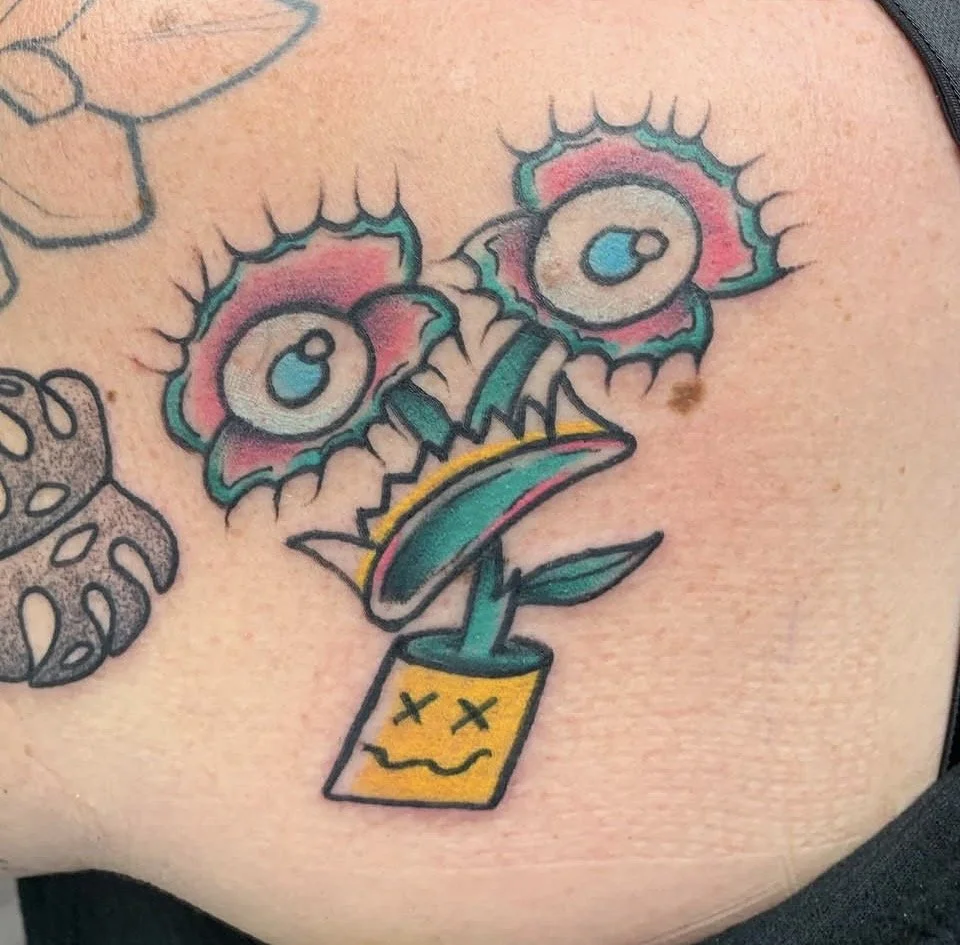 A tattoo of a cartoon face with a flowerpot for a head, featuring circular eyes with highlights, a mouth depicted as a smile, bright colors, and a bare-skin background.