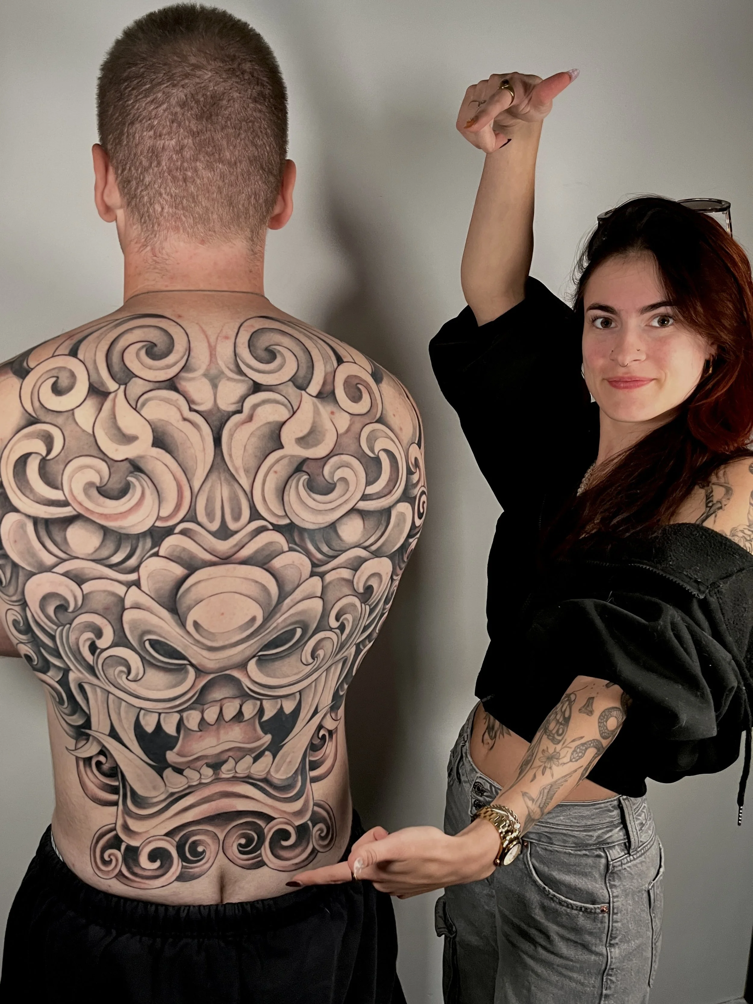 Une femme avec des tatouages pointant vers un énorme tatouage sur le dos d'un homme. Le tatouage représente un visage de créature mythologique ou de monstre bizarre et élaboré.
