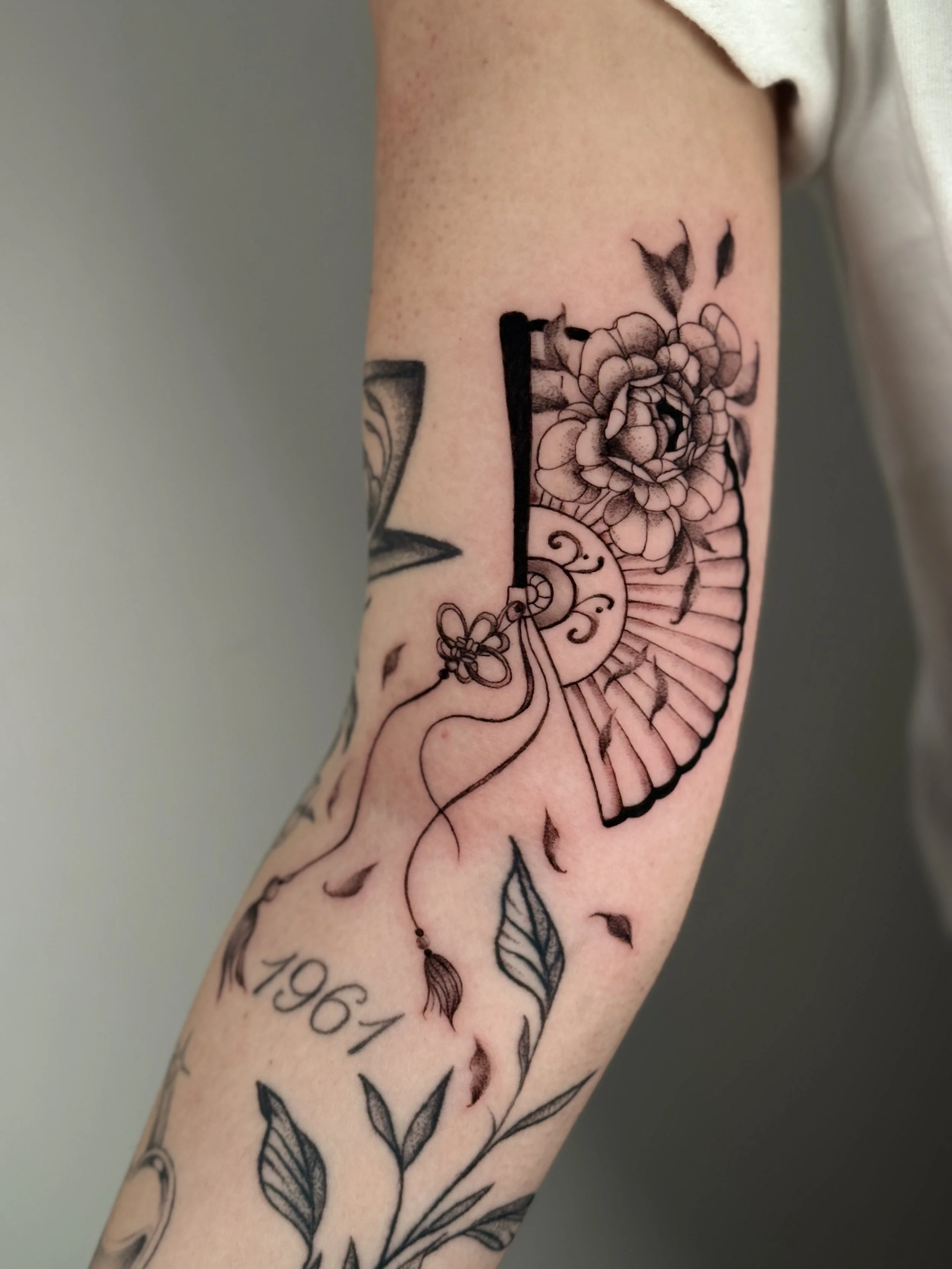 Black and gray tattoo of watch, flower, fan, leaves and 1961 date on arm