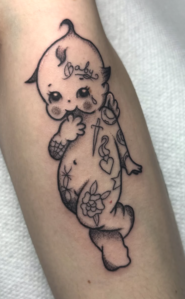 Tattoo of a baby mermaid with flowers, a heart, a pen, and the word "Baby" on her head.