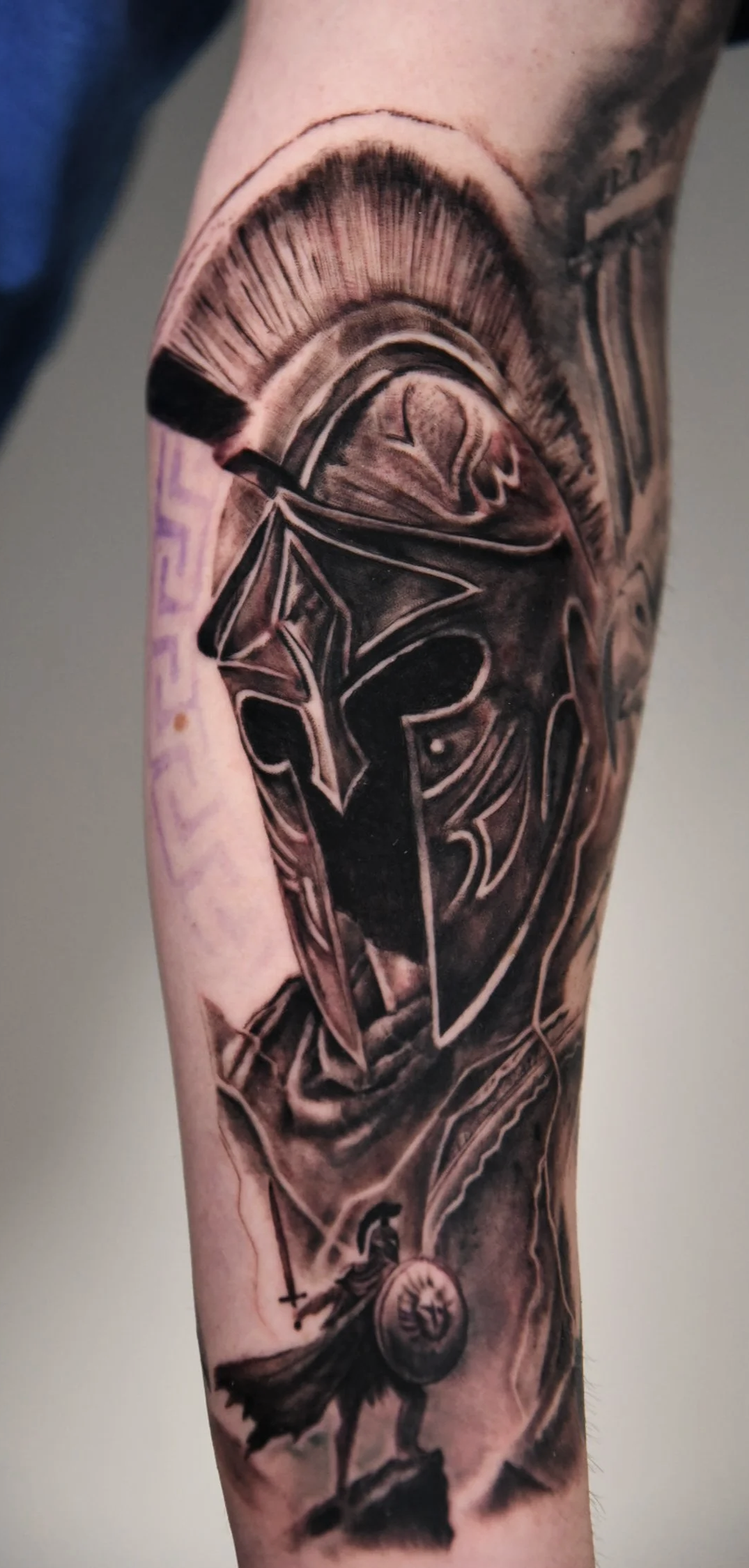 Black and gray tattoo of a military helmet with a spartan helmet in the center, and a warrior with a shield and sword at the bottom.