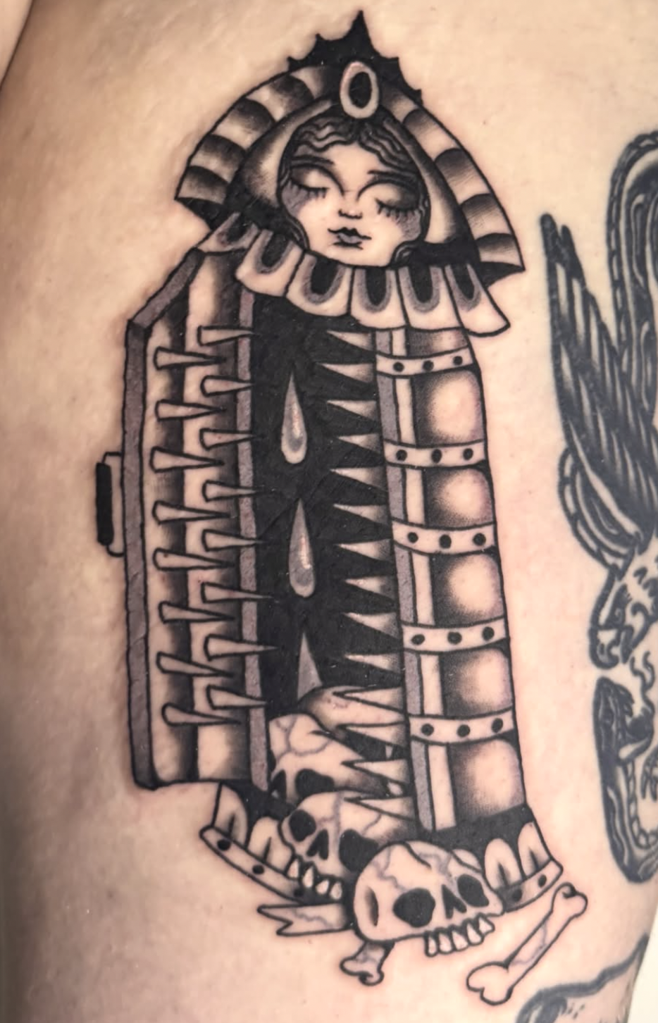 A black-and-white tattoo of a woman with a serene expression, surrounded by symbolic elements such as eyes, hearts, bones, and teardrops.