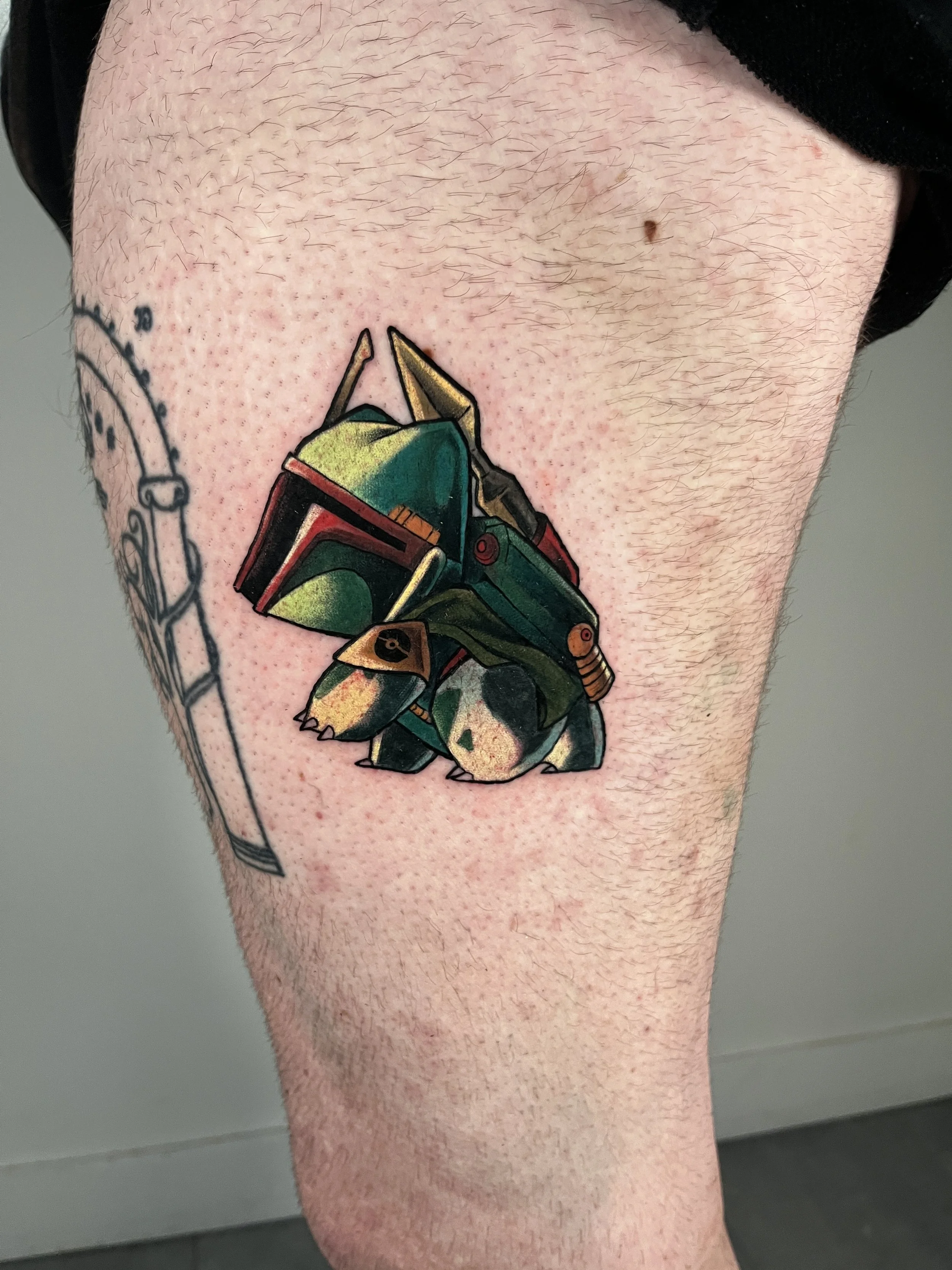 A colorful tattoo on a person's thigh depicting a Star Wars helmet—a green helmet with yellow and red details—in a cartoon style.