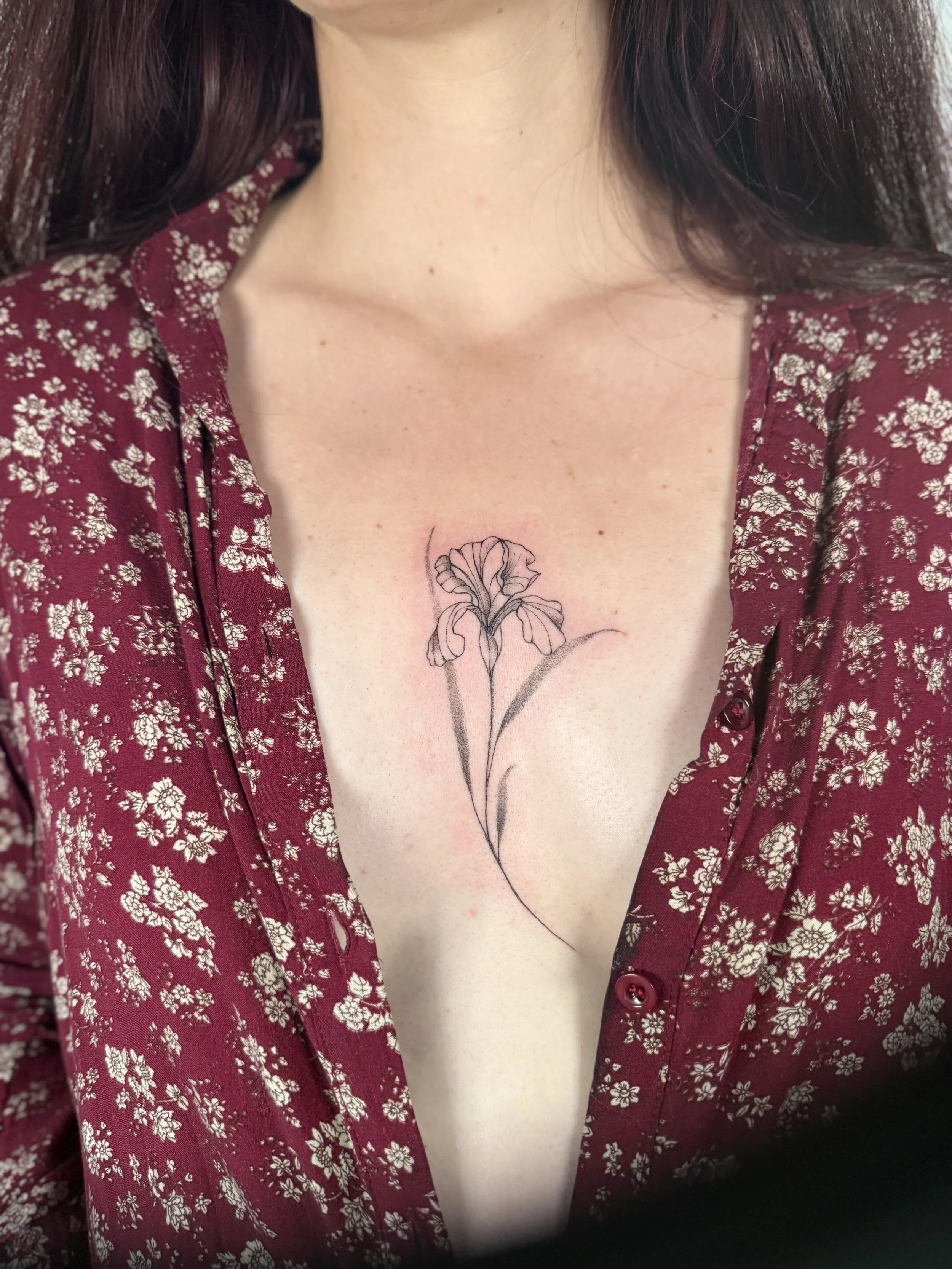 A woman wears a red shirt with small white flowers, open at the neckline, with a tattoo of a black flower on her chest.