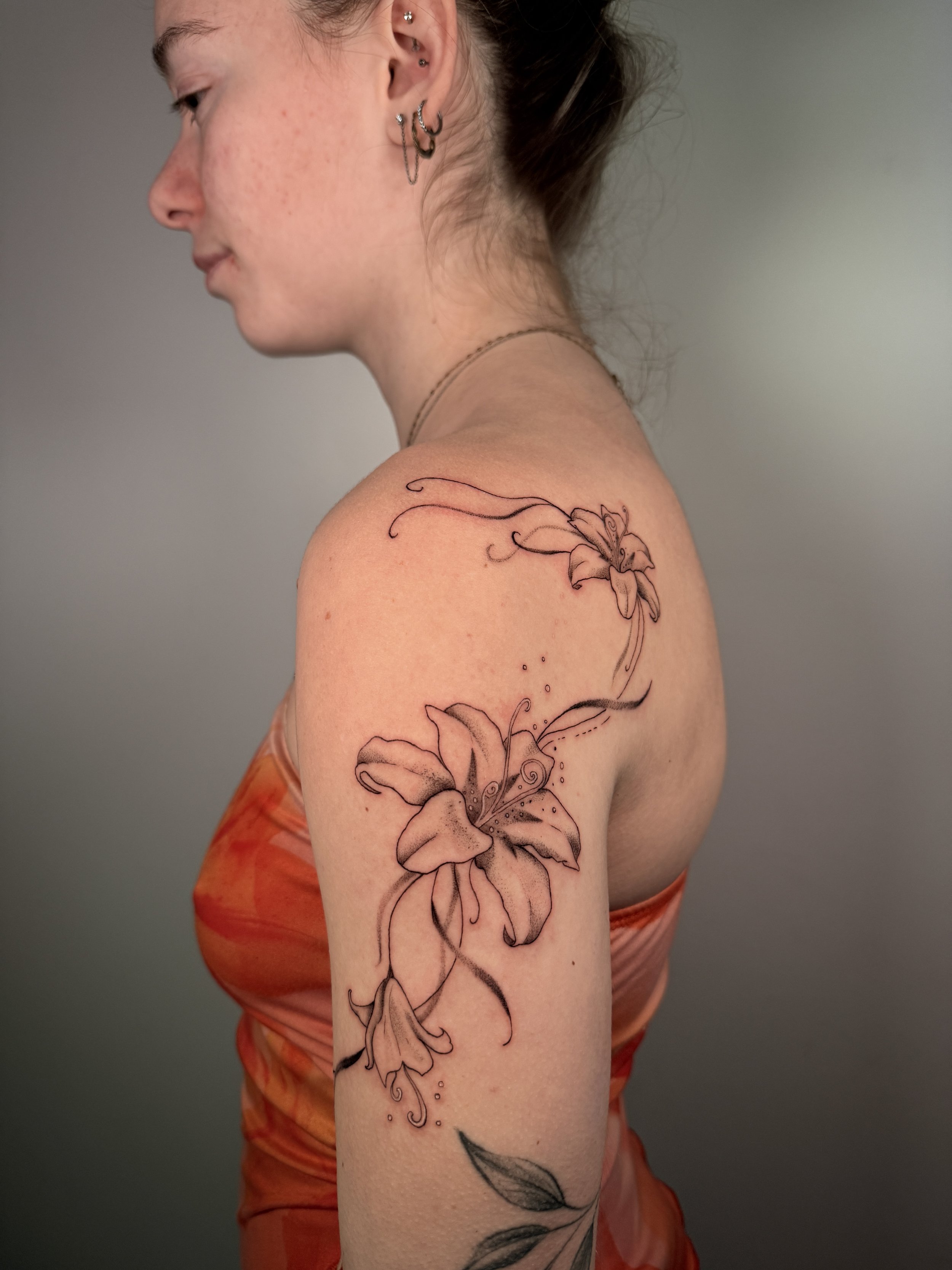 A young woman with several ear piercings and a black-and-white lily tattoo on her left upper arm.
