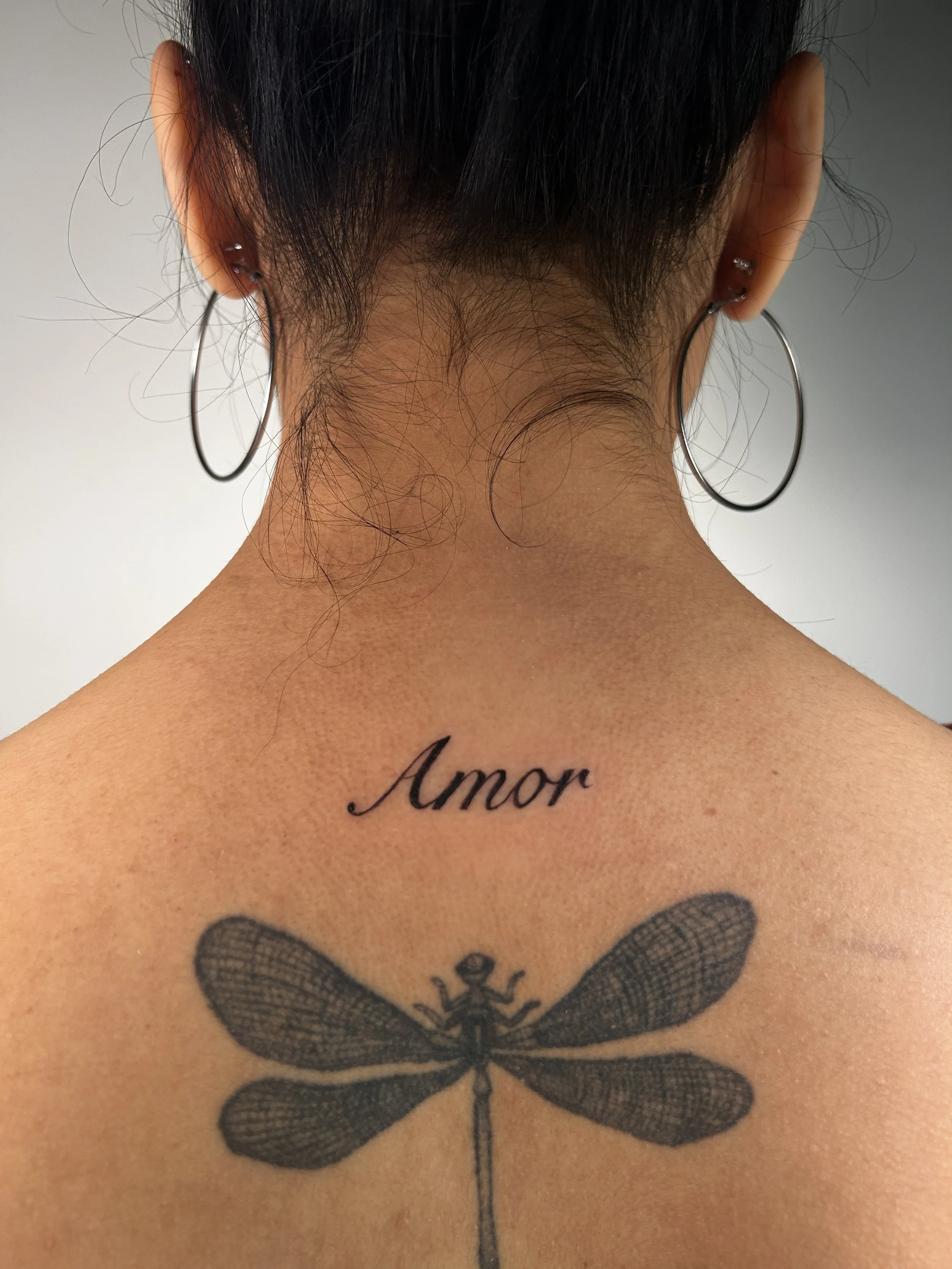 Tattoo of a dragonfly and the word 'Amor' written in cursive on the upper back of a woman, seen from behind, wearing circle earrings.