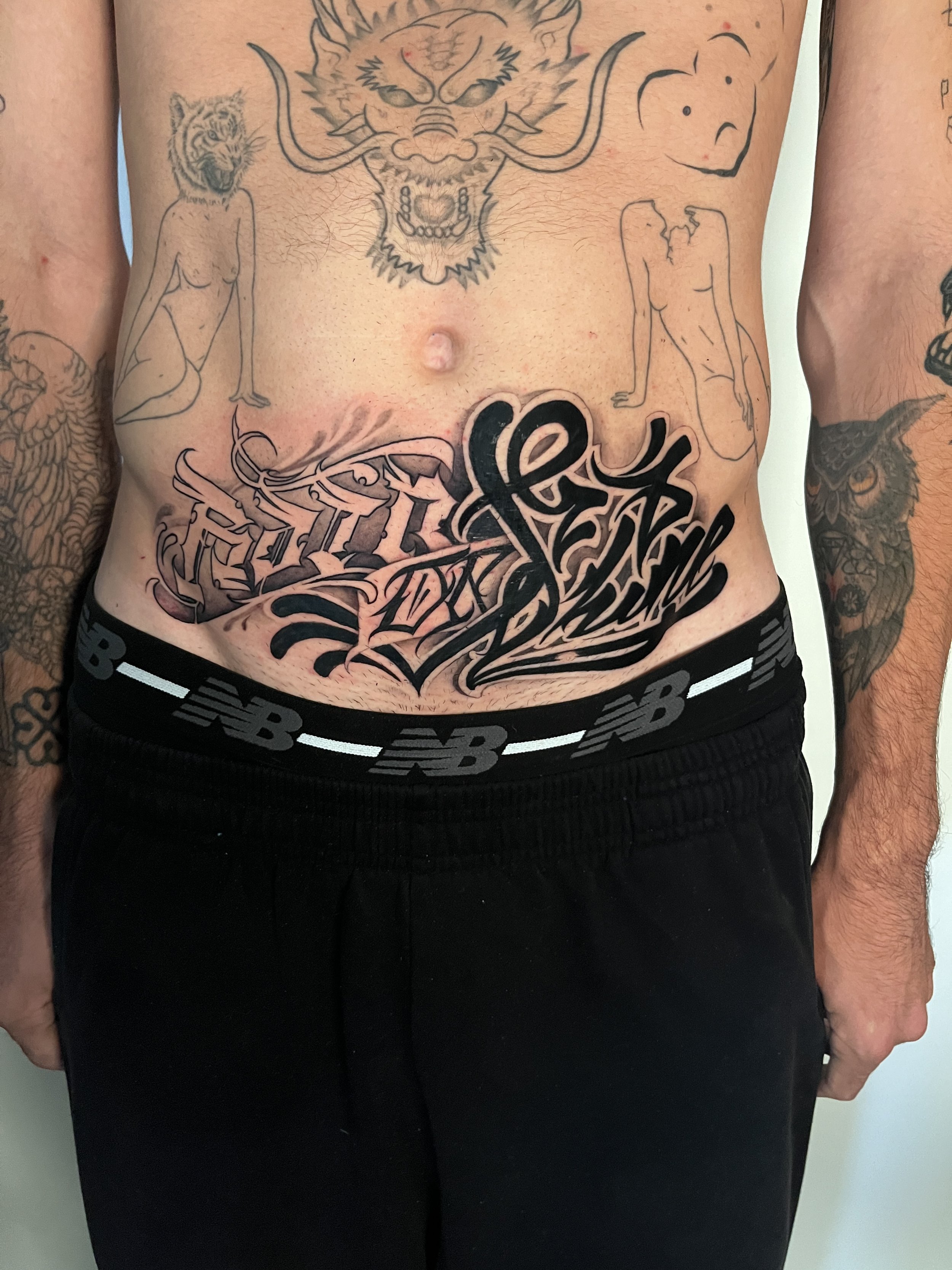 A man with several tattoos on his torso and arms, wearing black pants with a New Balance belt.