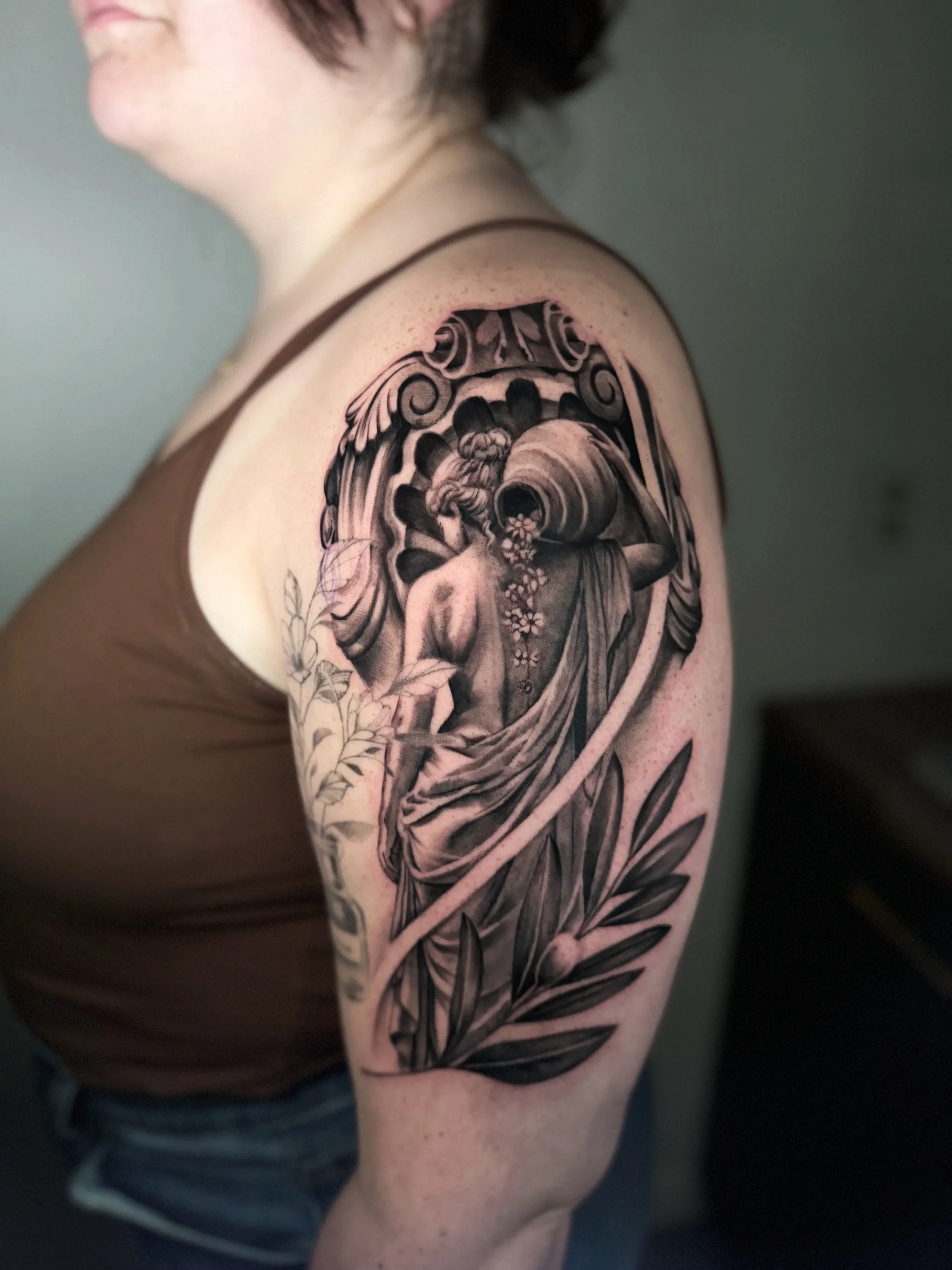 A grayscale tattoo on a person's shoulder depicting a scene featuring a female figure and an urn, surrounded by decorative patterns and leaves.