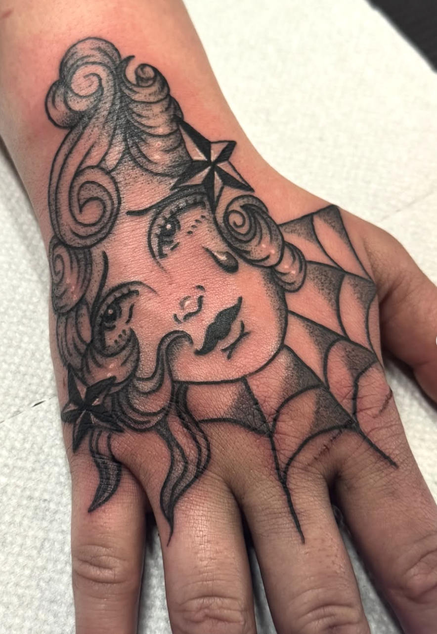 A black-and-gray tattoo of a woman with curly hair, wearing a dress with a spider-shaped pattern and a star on her forehead.