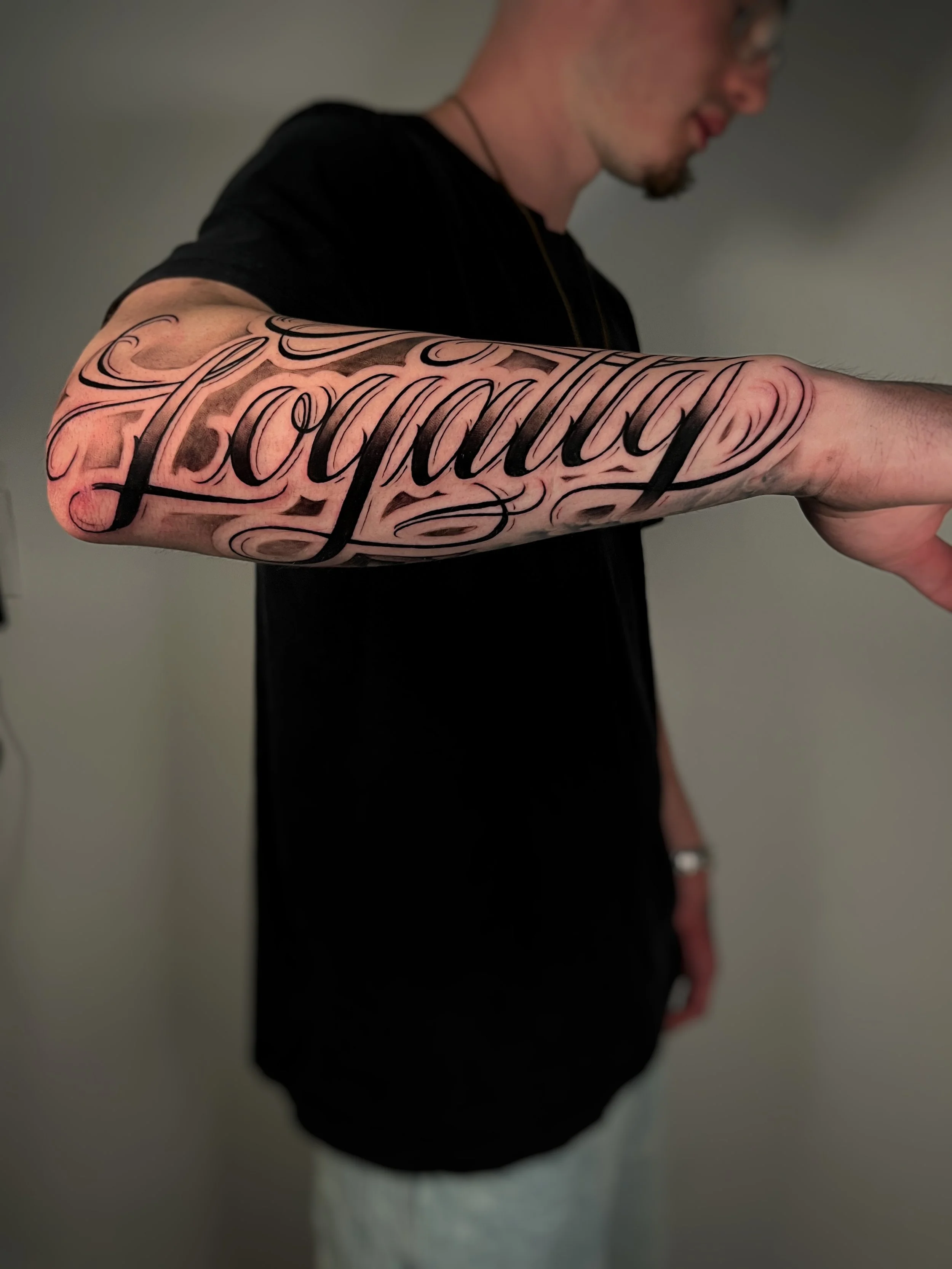 A script-style tattoo on the arm of a bearded man wearing a black T-shirt.