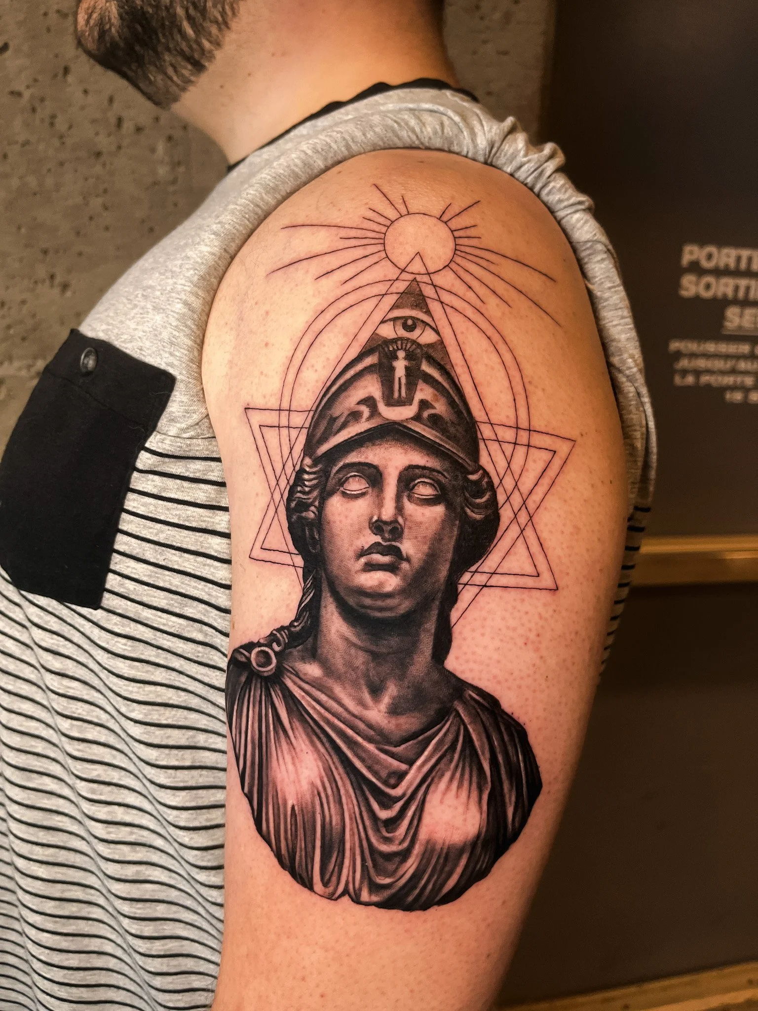 Tattoo of a female statue with helmet, surrounded by geometric shapes and a sun at the top.