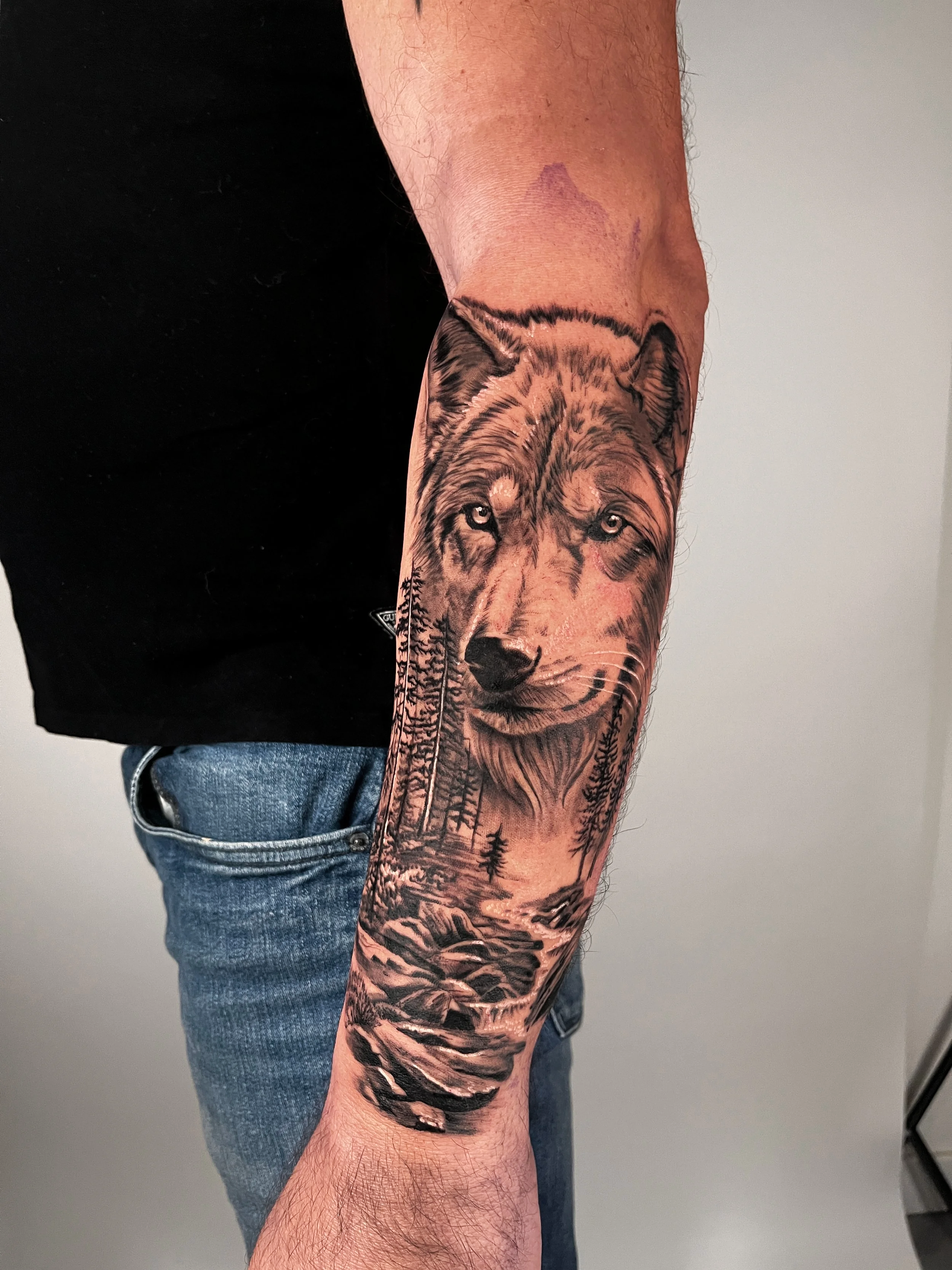 Realistic tattoo of a wolf with a forest and a river on a person's arm.