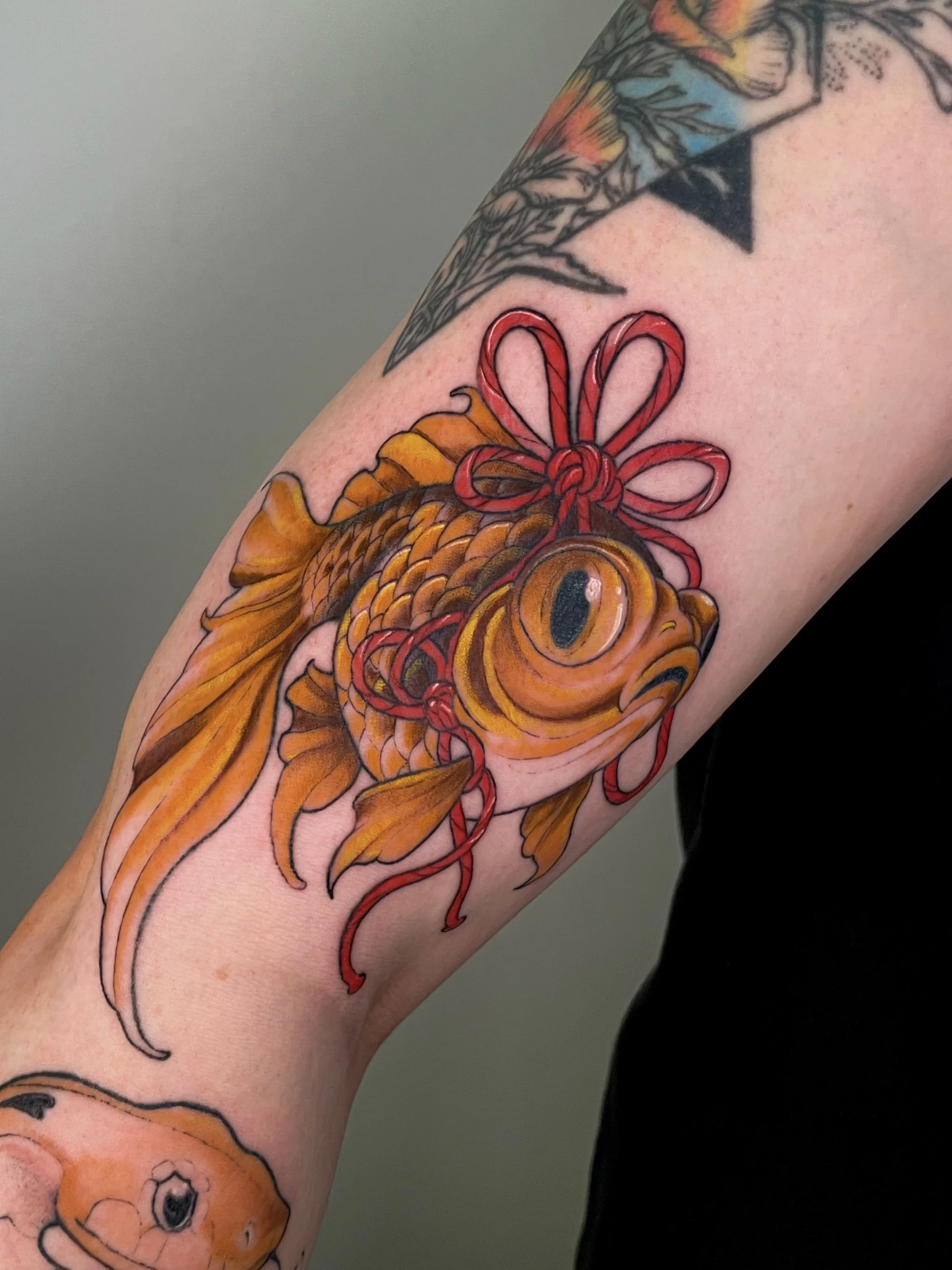 Tattoo of a goldfish with a wide eye and flowing tail, with a red bow at the top, on a person's arm.