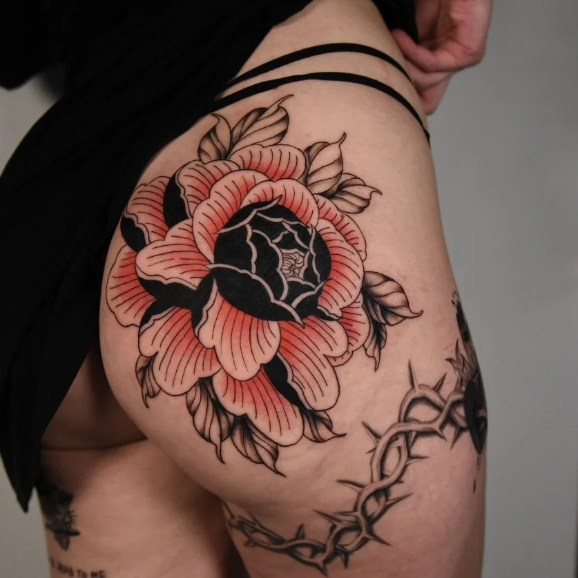 Tatouage d'une grande fleur rose avec des feuilles et des détails noirs sur la cuisse d'une personne.