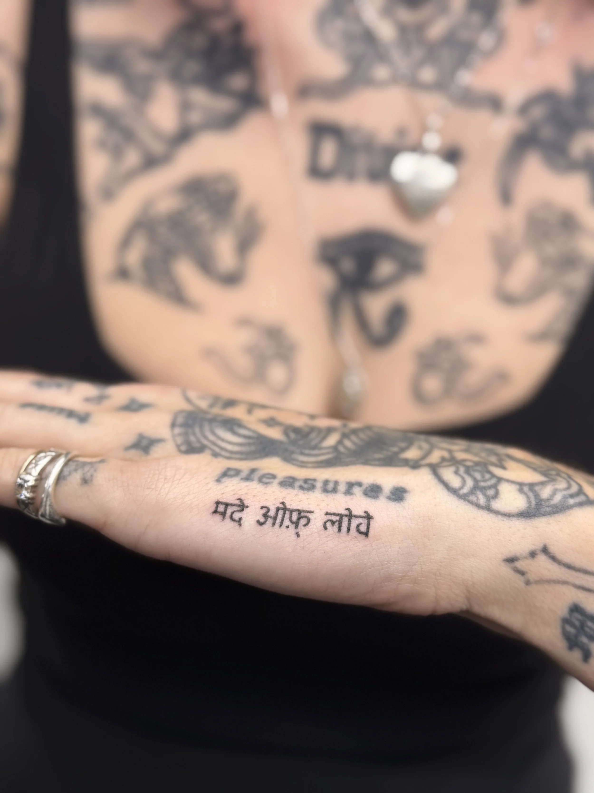 Right hand tattooed with the word 'pleasure' in English and a sentence in Hindi. In the background, a person with numerous facial tattoos.