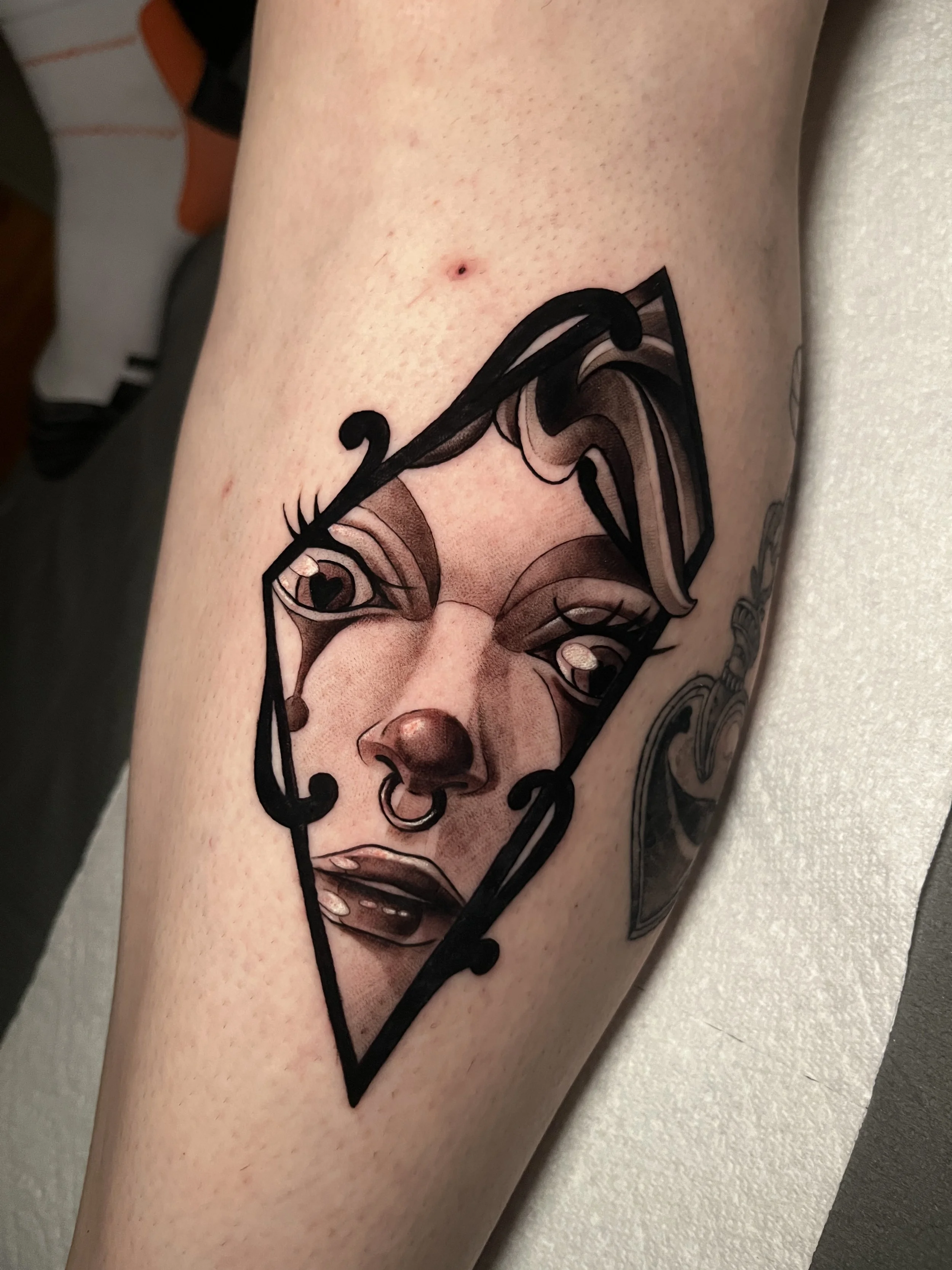 Realistic tattoo of a woman's face with dark makeup, an earring at the nostril, framed by a black geometric shape on the skin