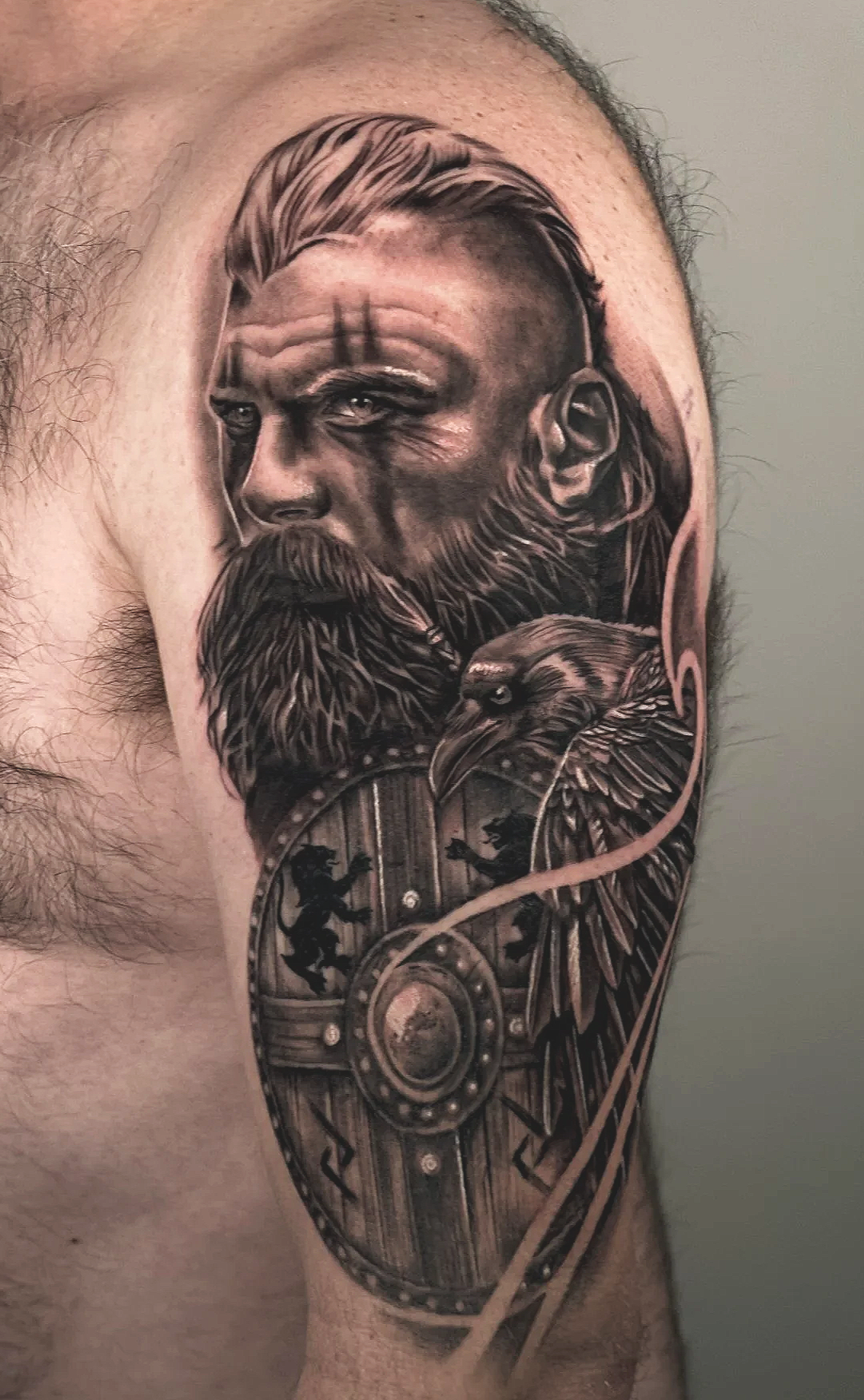 Realistic tattoo depicting a bearded man's face with sophisticated hair, accompanied by an eagle and a Viking shield roundel adorned with engraved lions.