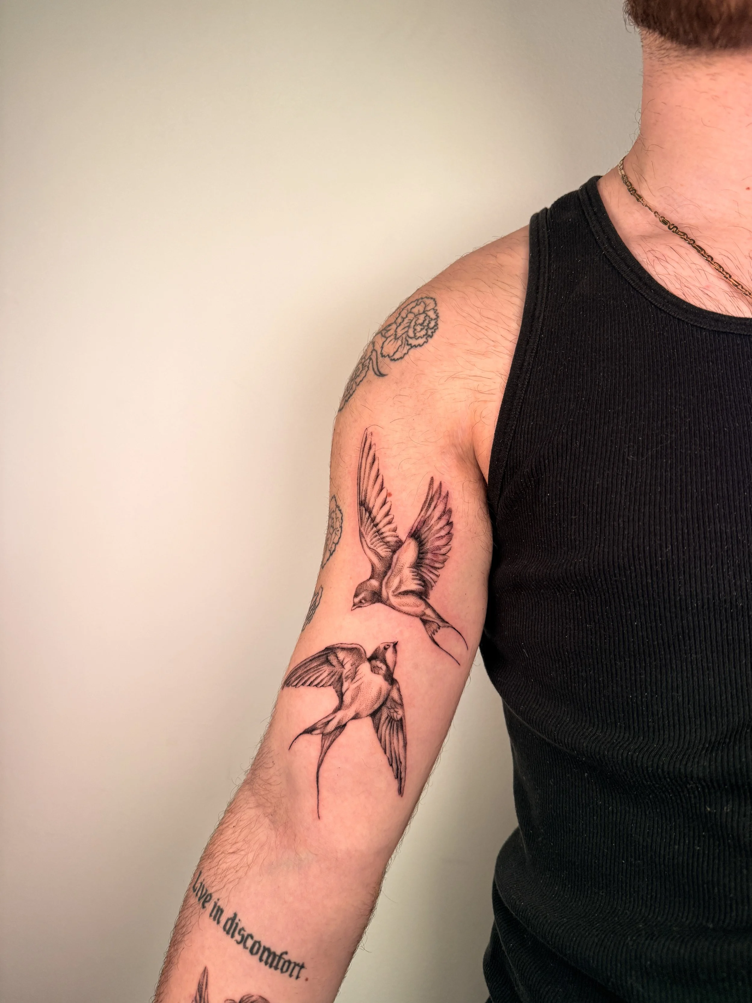 A man with a tattoo of birds on his right arm and a necklace is wearing a black T-shirt and standing against a beige wall.