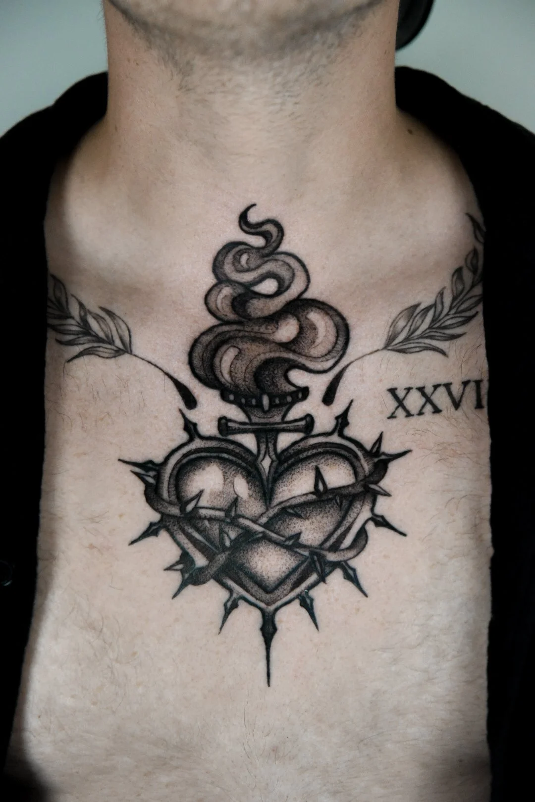 Tattoo on the chest of a heart surrounded by brambles, with a flame above and leaves on either side.
