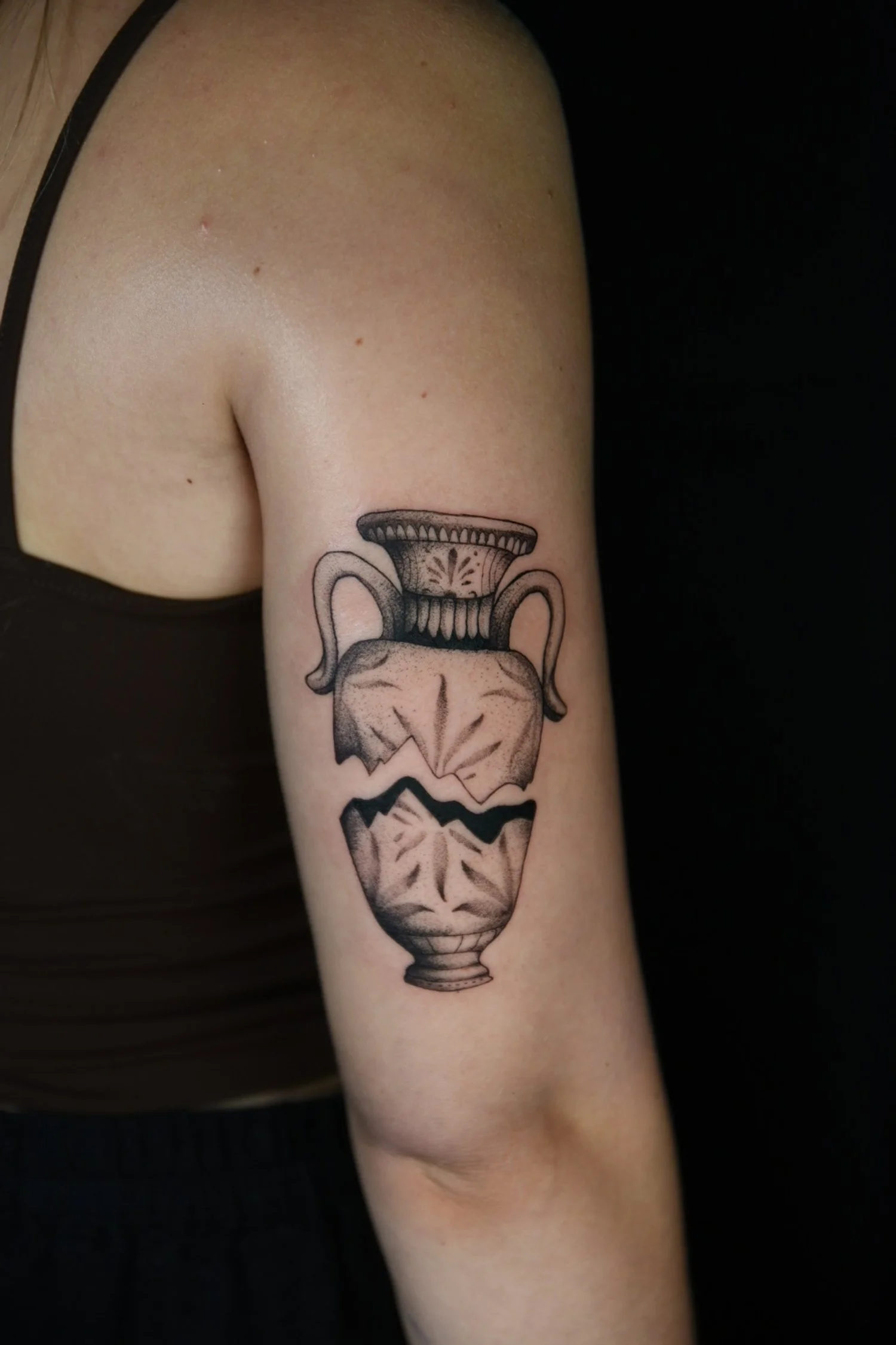 Black and gray tattoo of an antique dish on the upper arm, depicting a broken urn or vase.