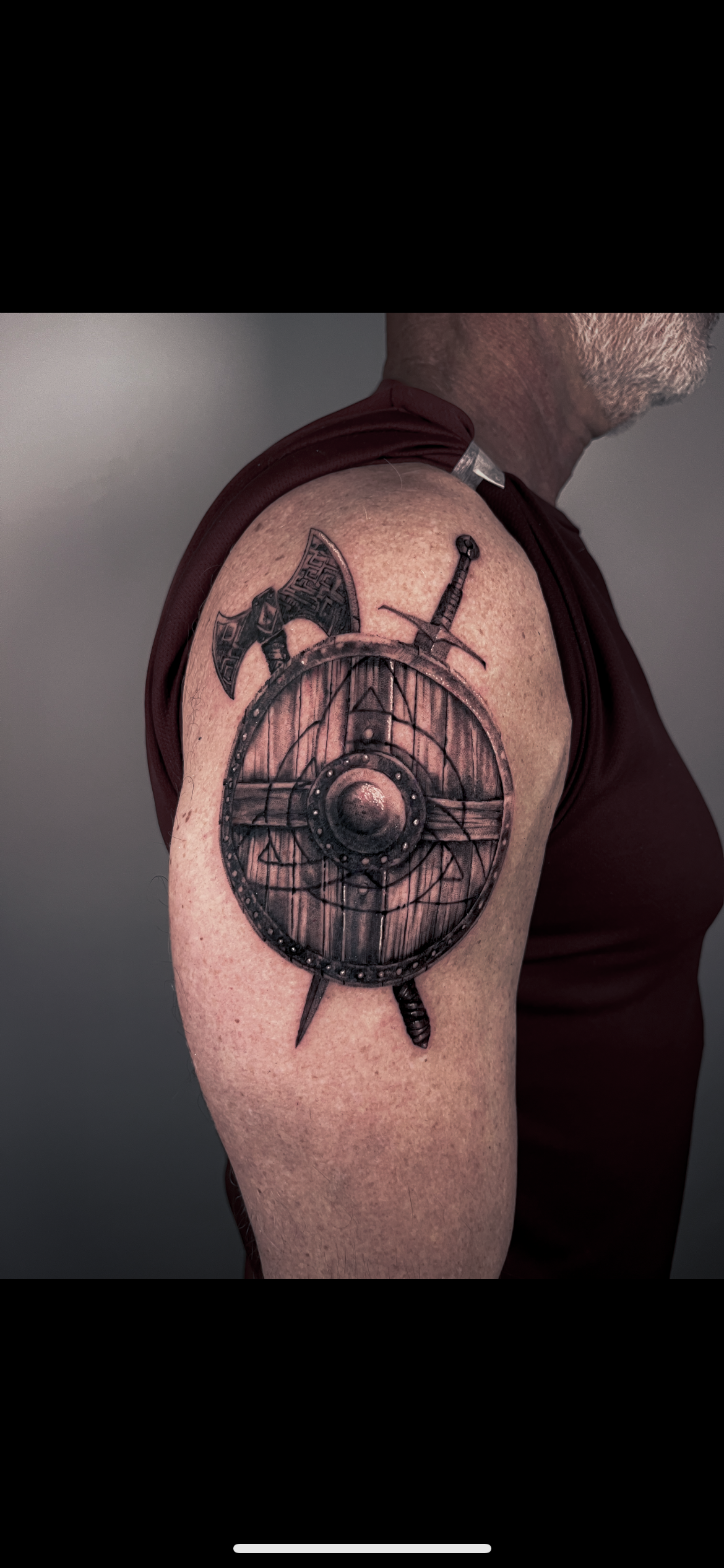 Tattoo of a wooden shield with crossed axe and sword on a man's arm.