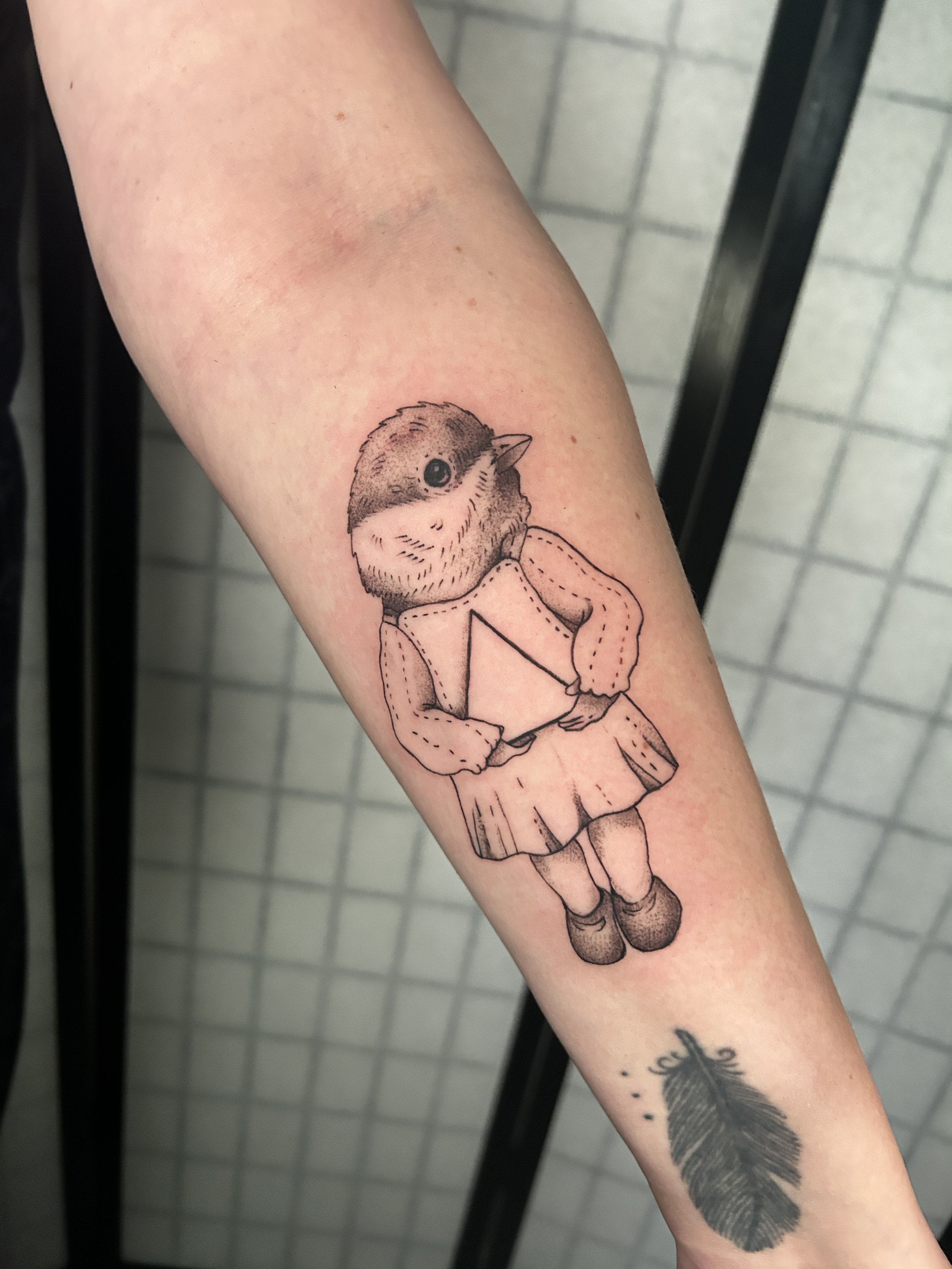 Tattoo of a small anthropomorphic bird wearing a dress and holding a book.