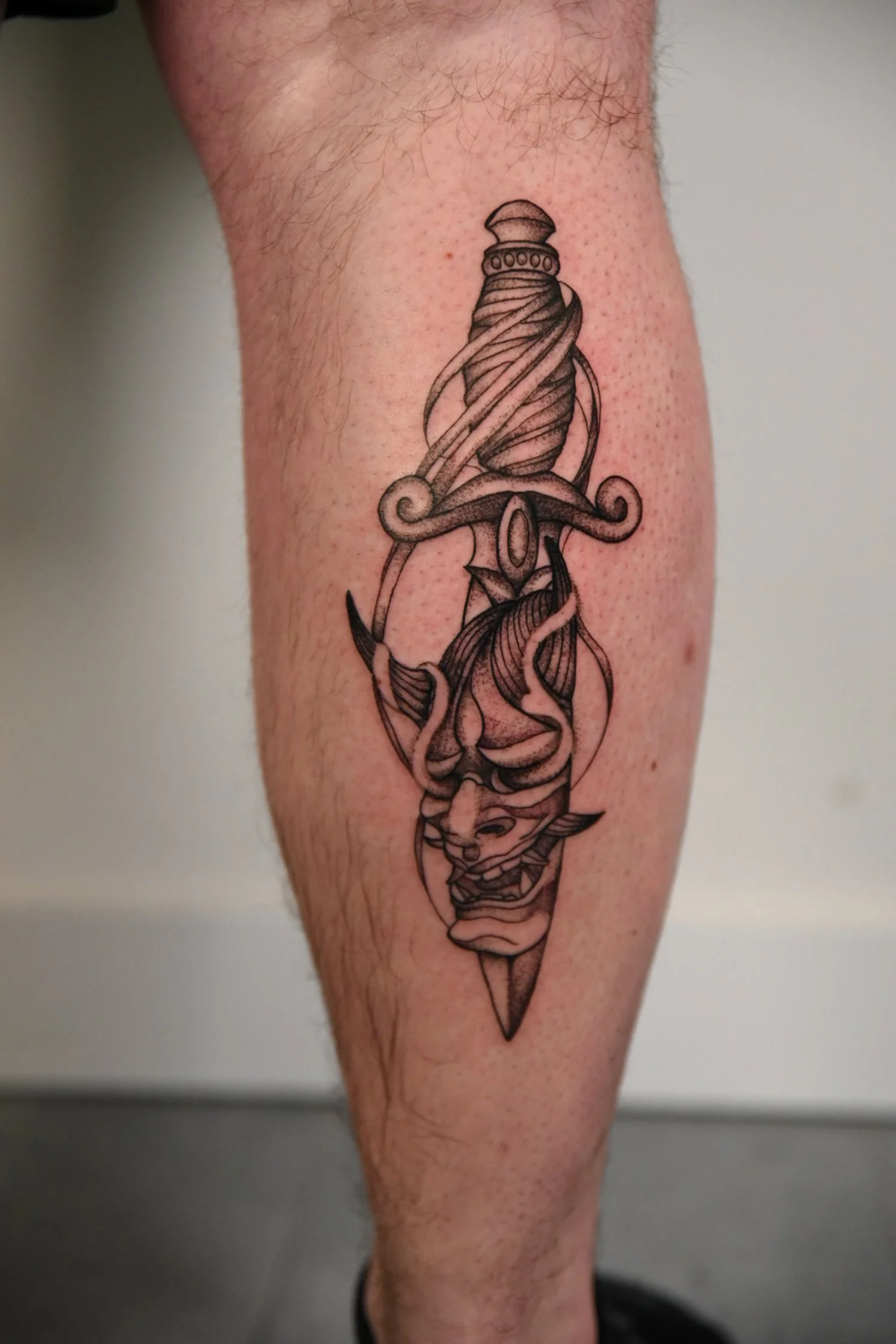 Tatouage noir et gris représentant une arme, une tête de démon ou de créature mythologique, et des éléments de décoration dans un style traditionnel.