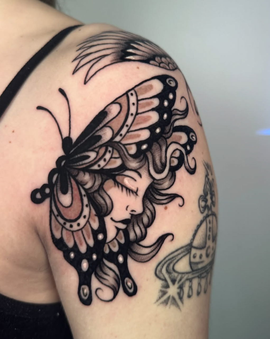 A tattoo of a woman with butterfly wings on her shoulder, stylized in black and white with intricate details.