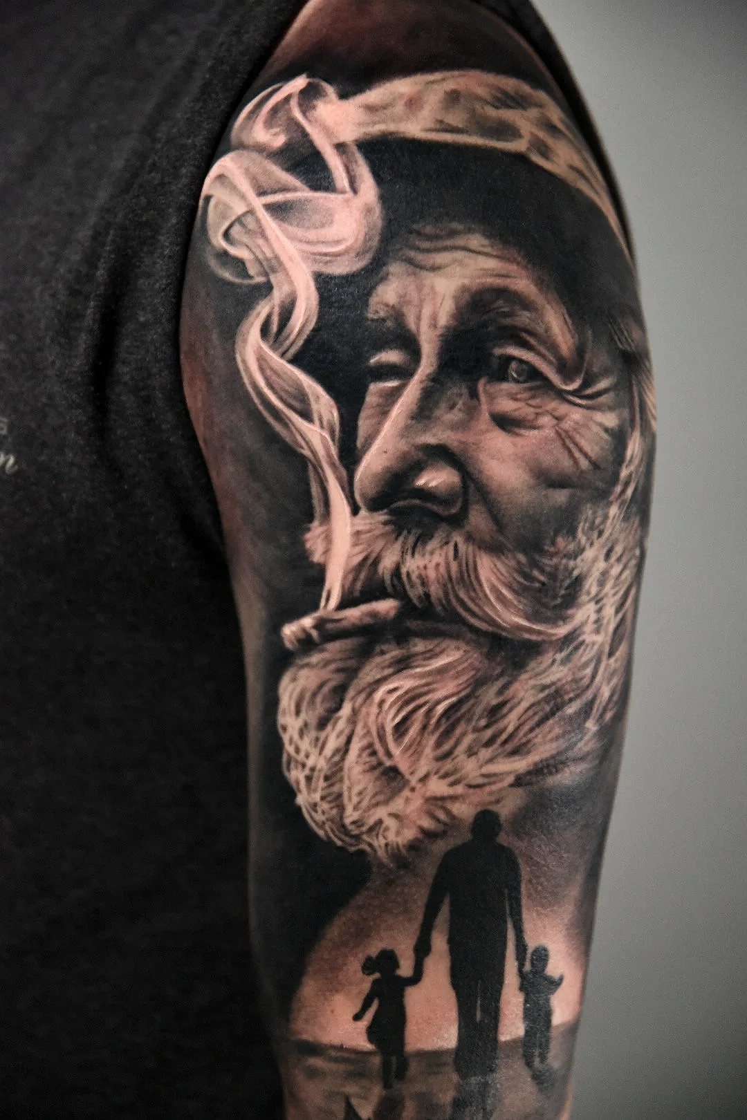 Black and white tattoo depicting an elderly man with a long beard and pipe, a silhouette of adults and children holding hands, with smoke rising into the air.