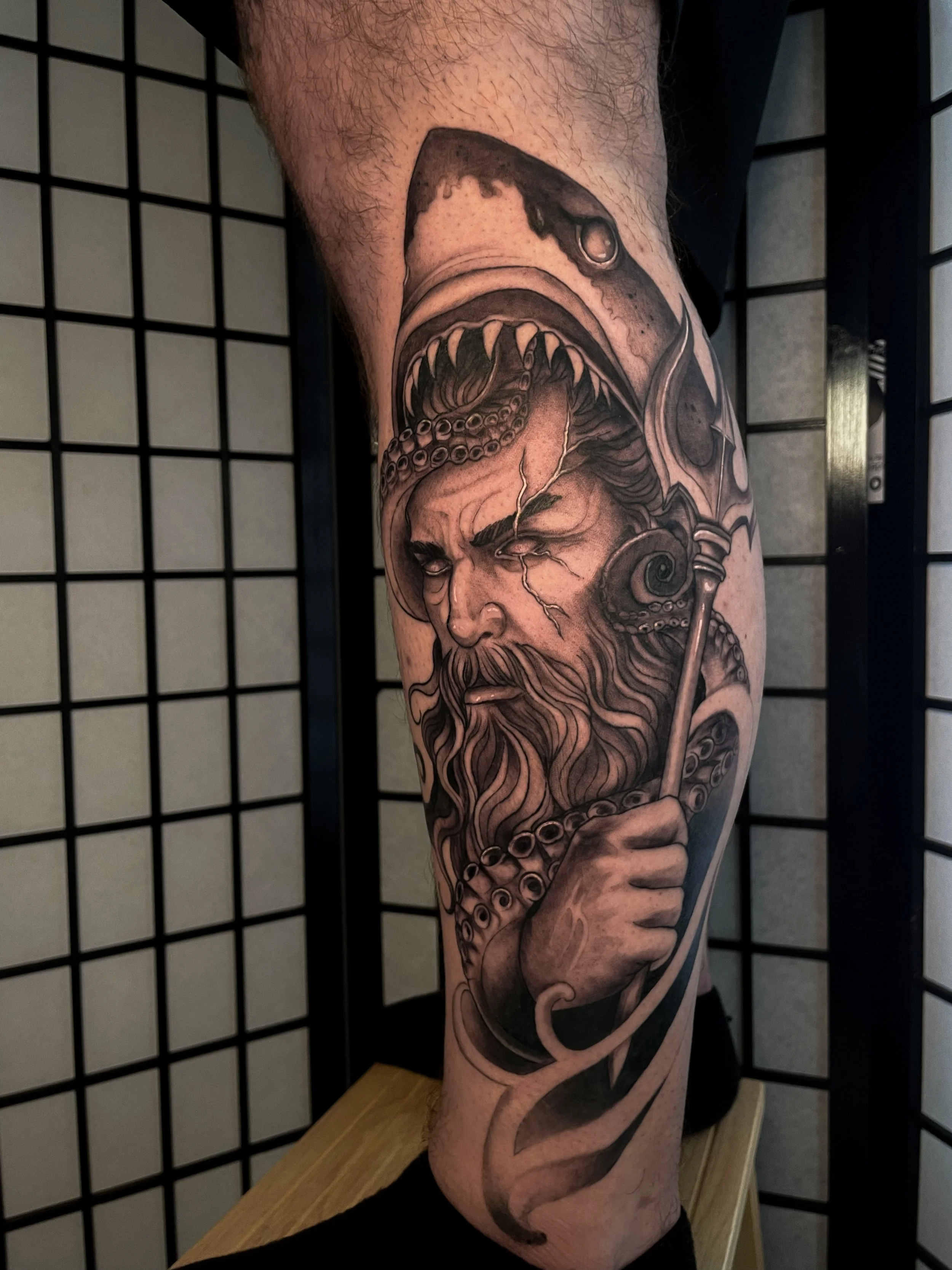Black and gray tattoo of a bearded man with a serious expression, holding a spear, with a mythology or fantasy-style face, including a fish or sea monster above.