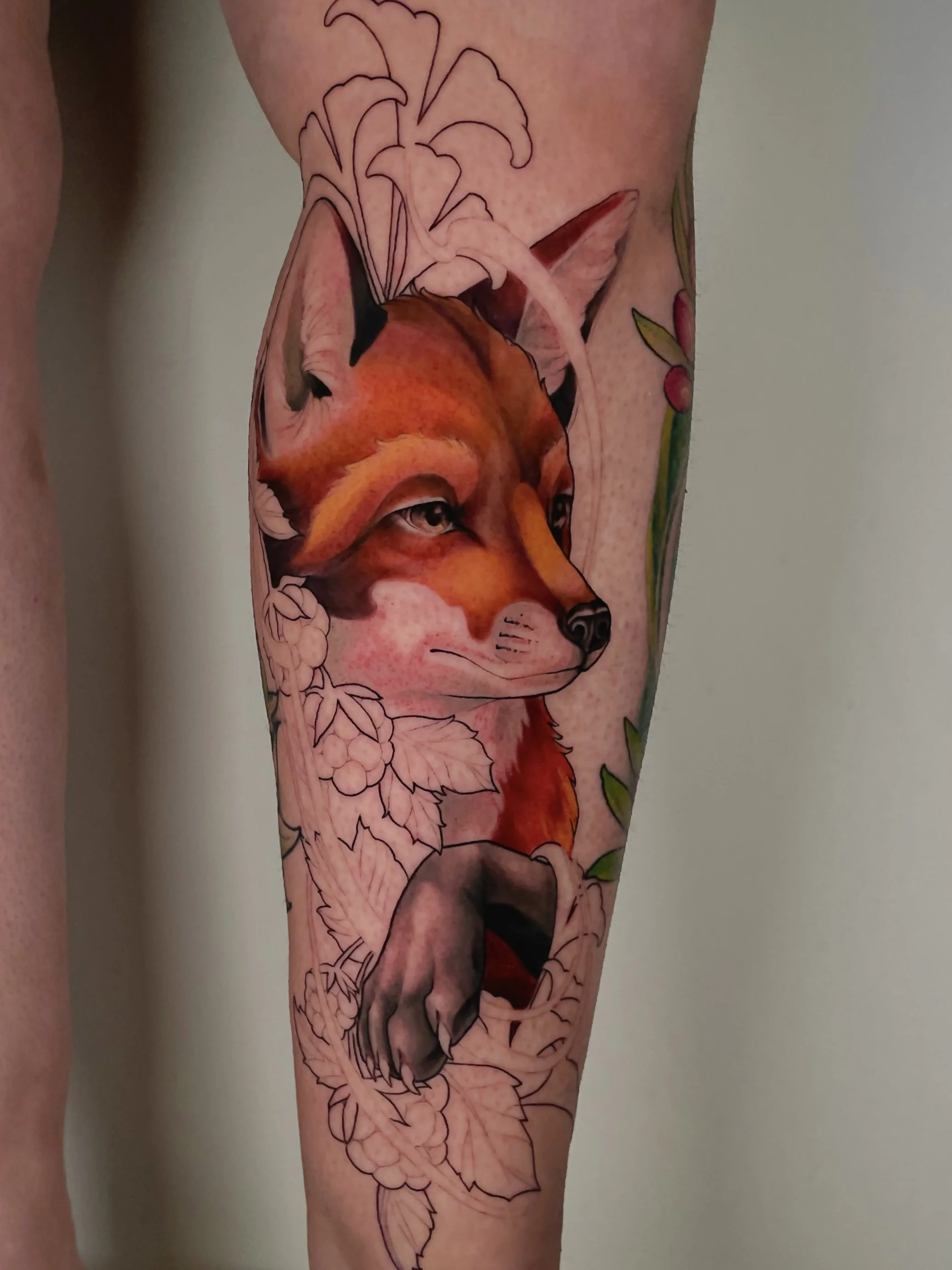 Tatouage d'un renard en partie coloré et en partie esquissé, avec des fleurs et feuillages en arrière-plan.