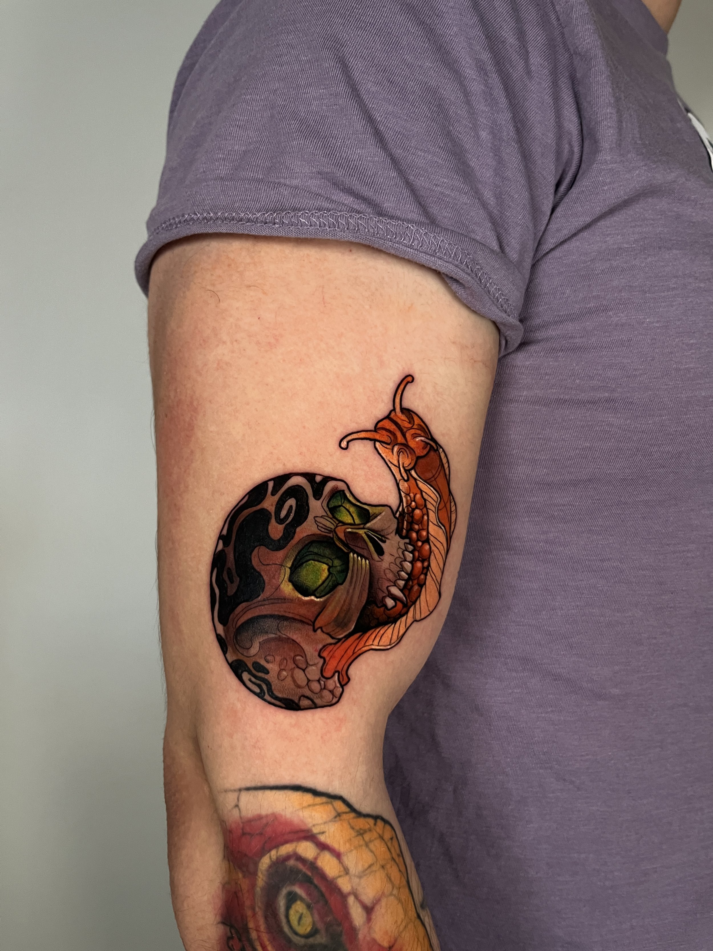 An arm with a colorful tattoo depicting a cannabis plant and an insect, belonging to a person wearing a purple T-shirt.