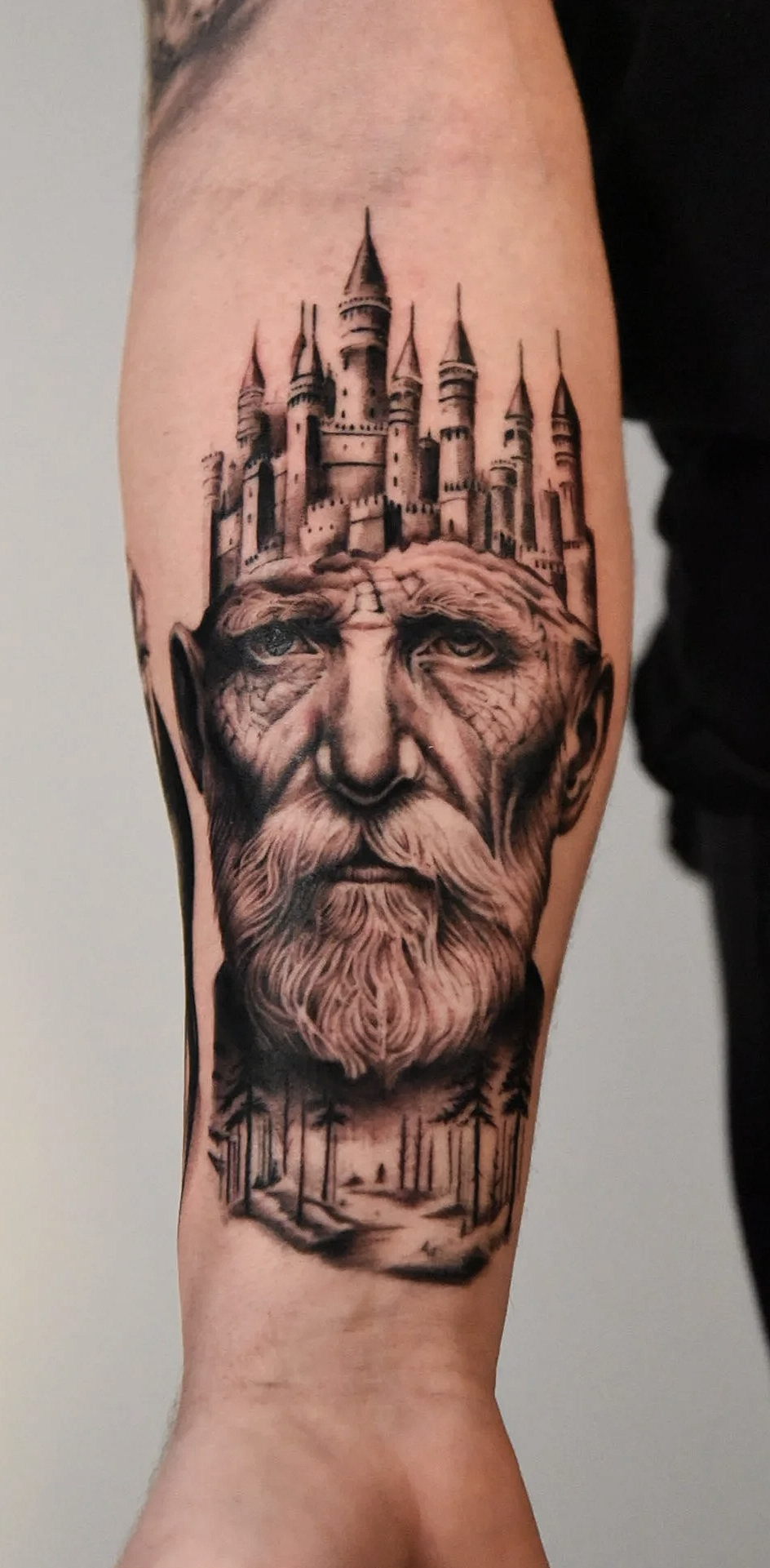 Tattoo depicting an old man's face with a long beard, with a medieval castle on his head, and a forest below.