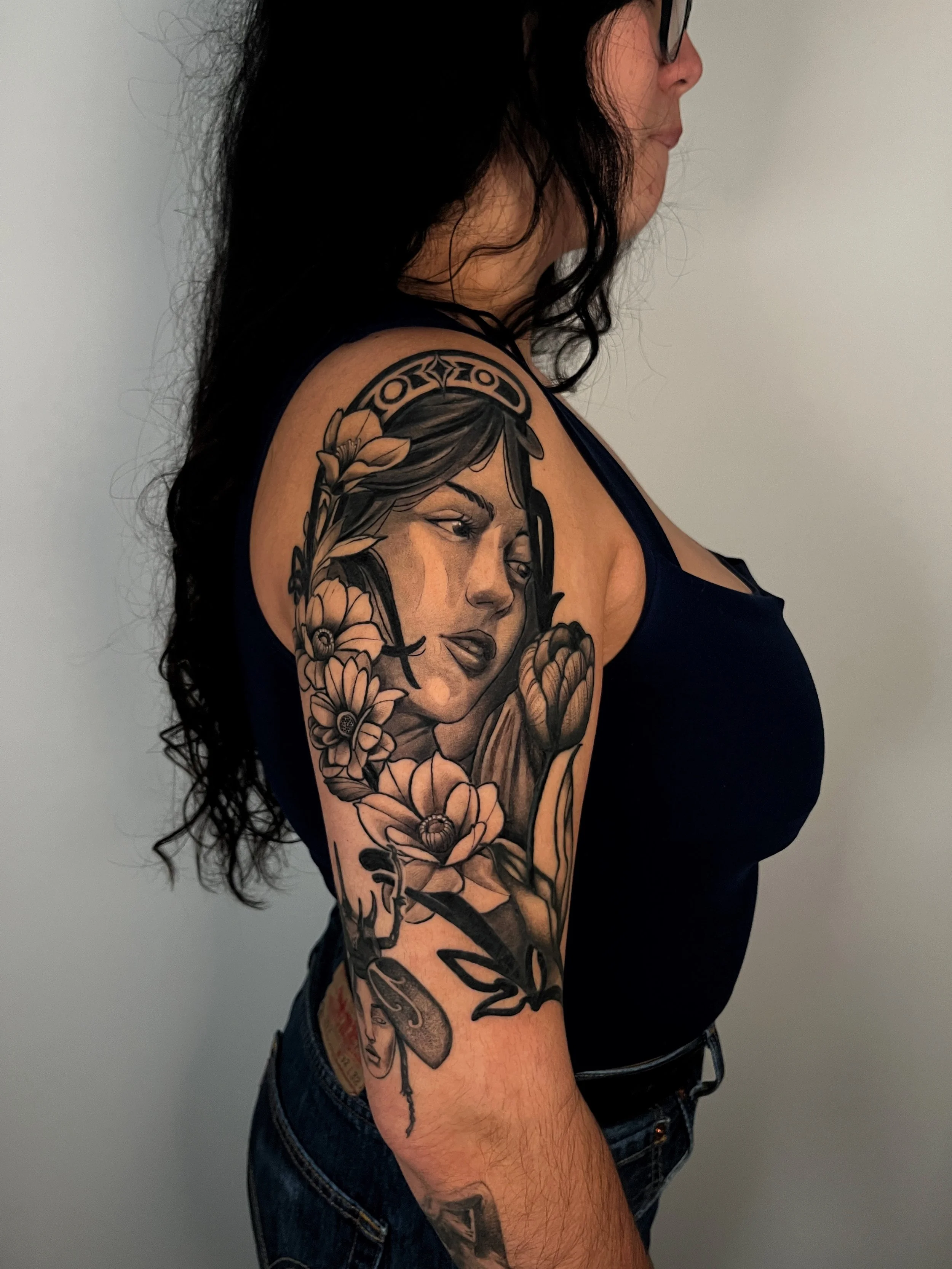 Tattoo of a female face surrounded by flowers and decorative elements on the arm of a woman with black hair and wearing a black top