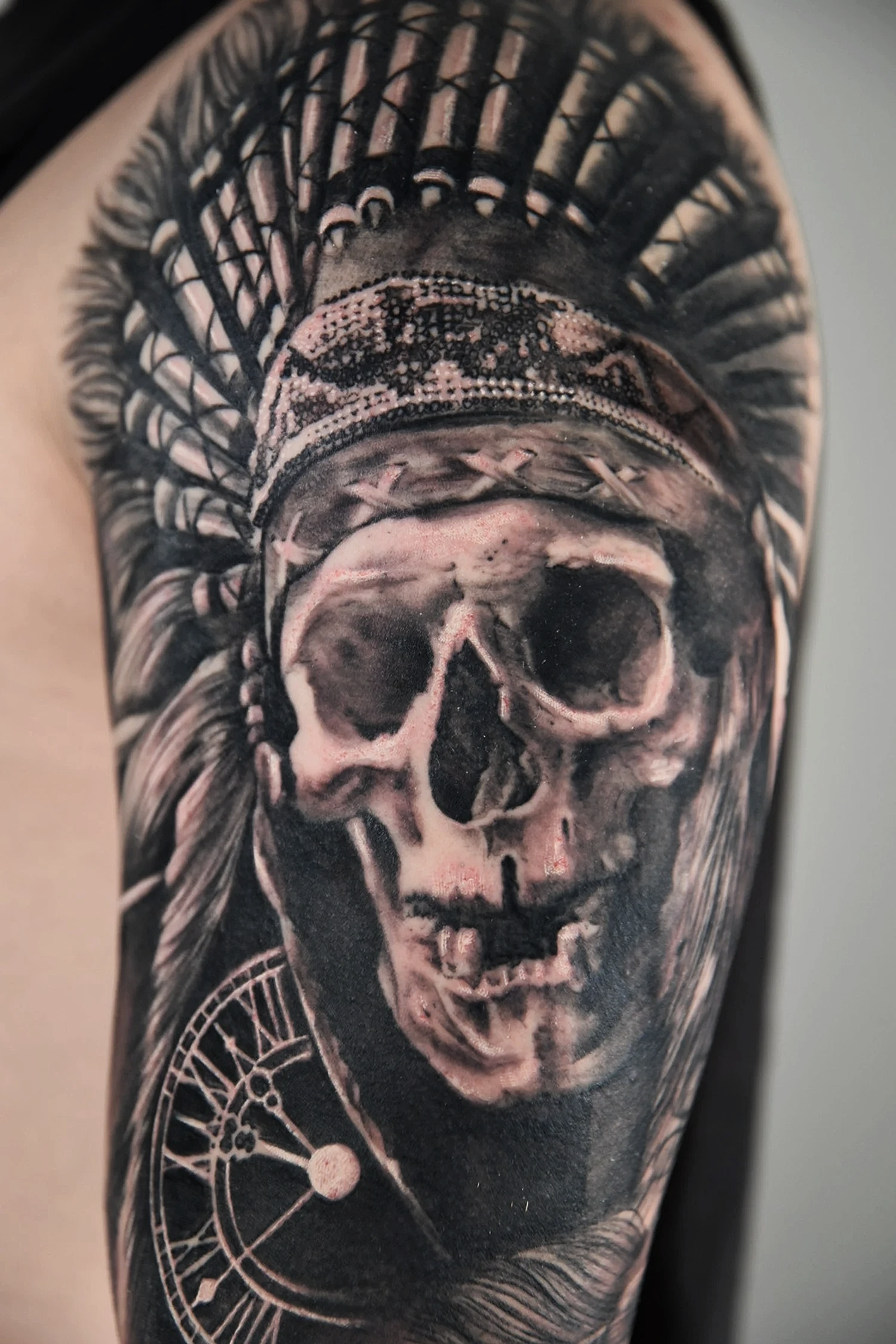 Black and white tattoo featuring a skull with Indian headdress, watch circle and feathers.
