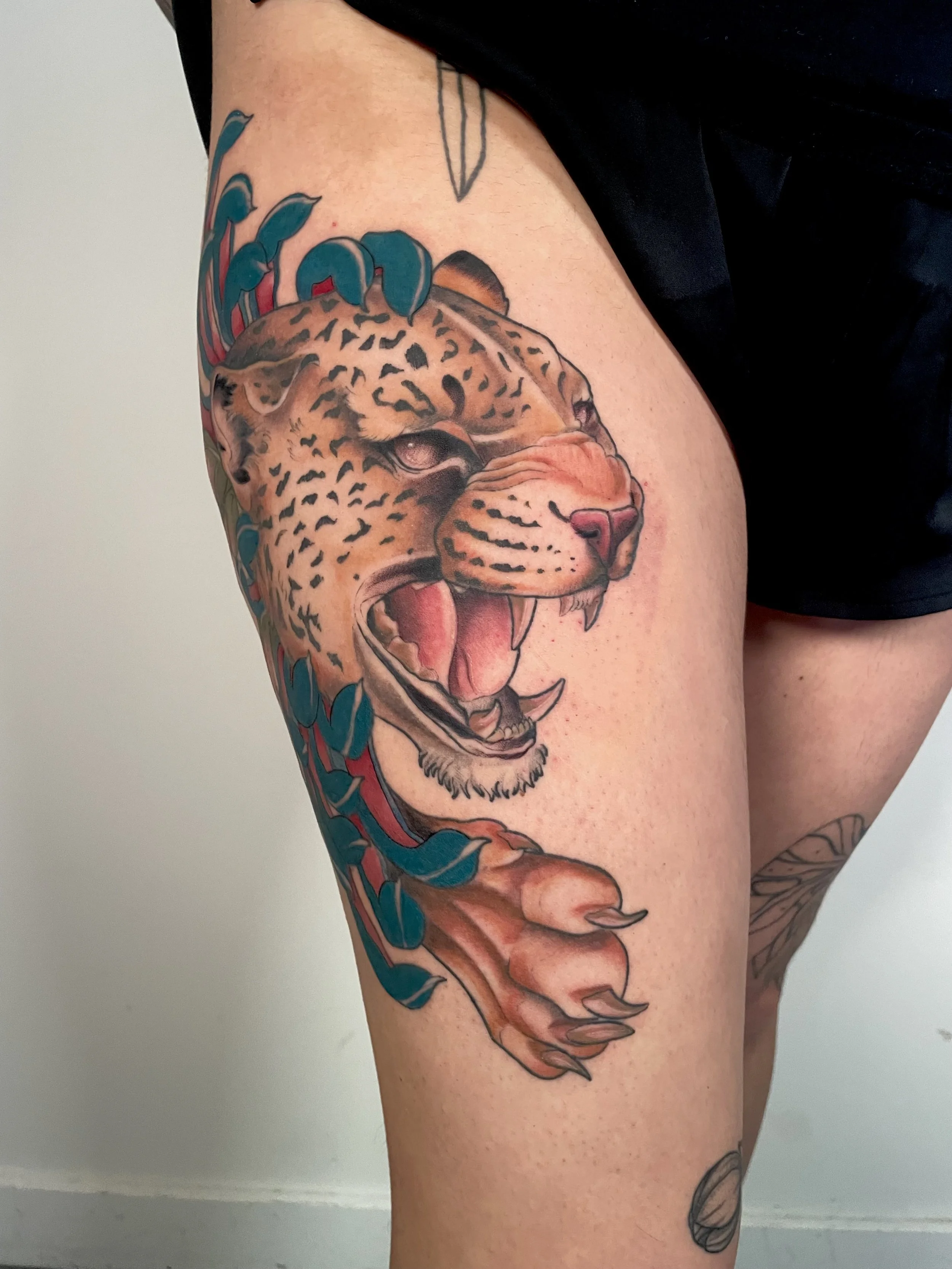 Tattoo of a roaring jaguar on the thigh, with blue leaves and colorful details.
