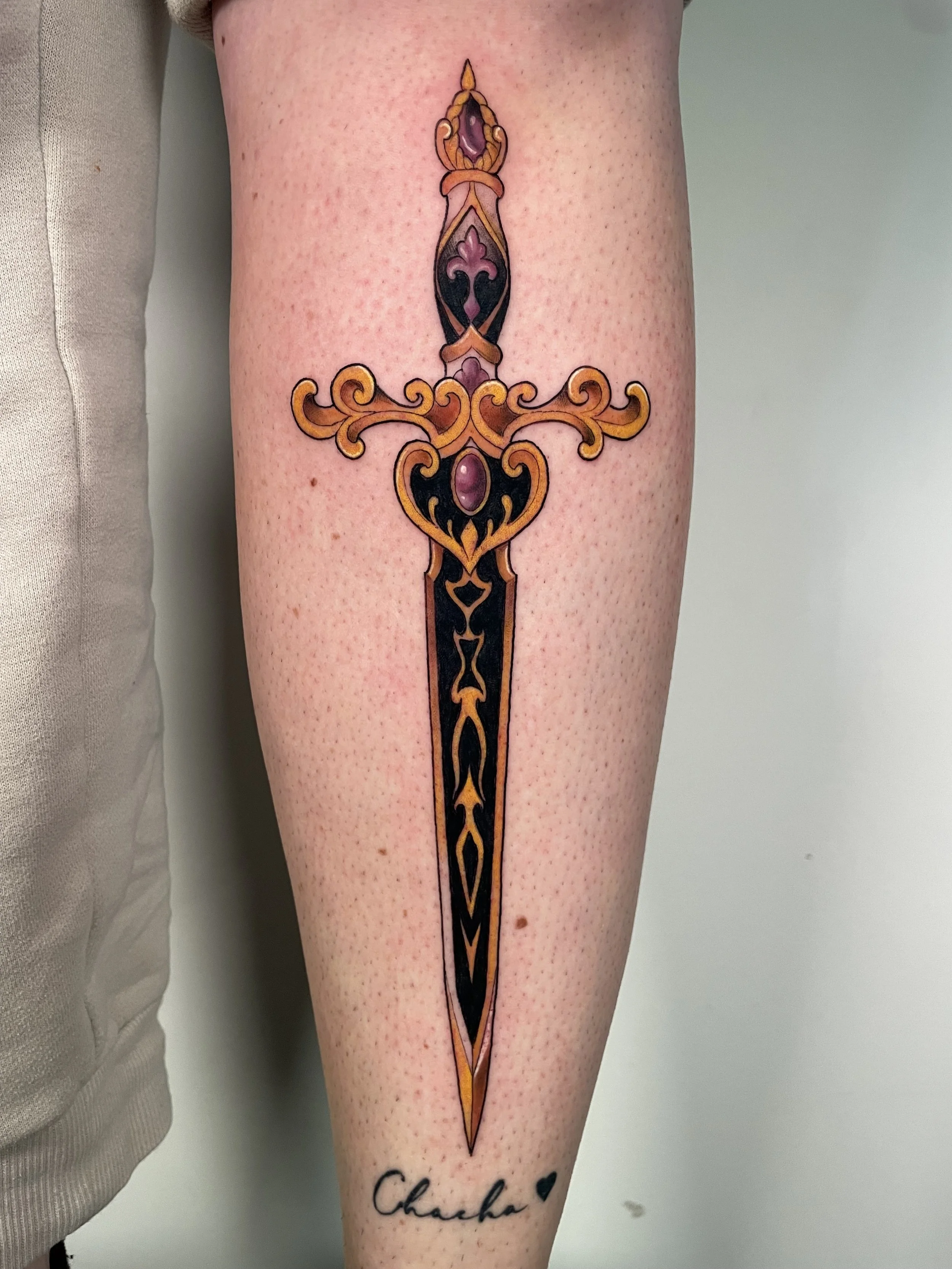 Tattoo of a sword with ornamental details and purple stones on the leg. Beneath the tattoo is a first name, "Chacha", written in cursive.