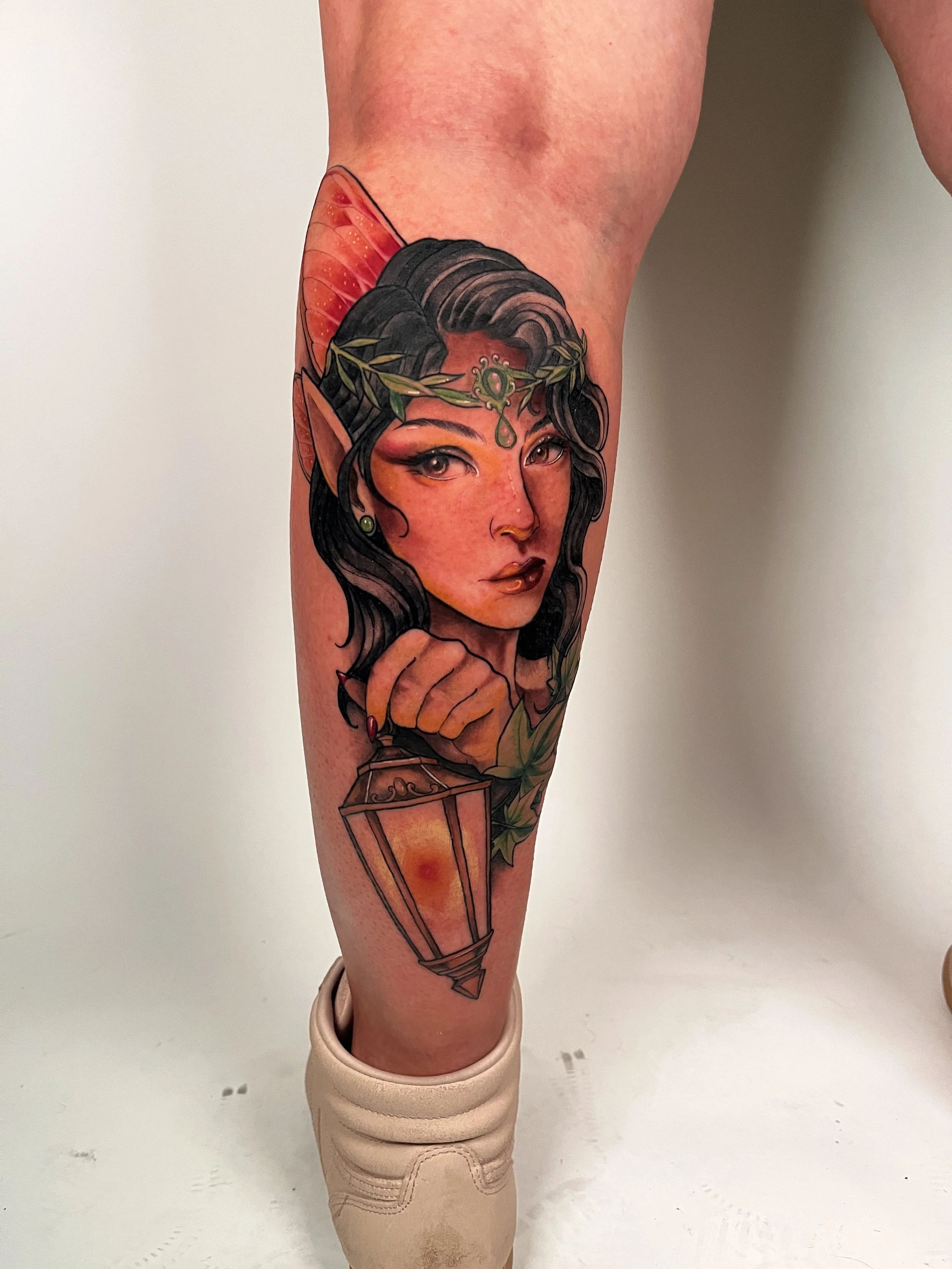Tatouage d'une elfe avec une couronne de feuilles, une lanterne, et des ailes rouges sur la jambe
