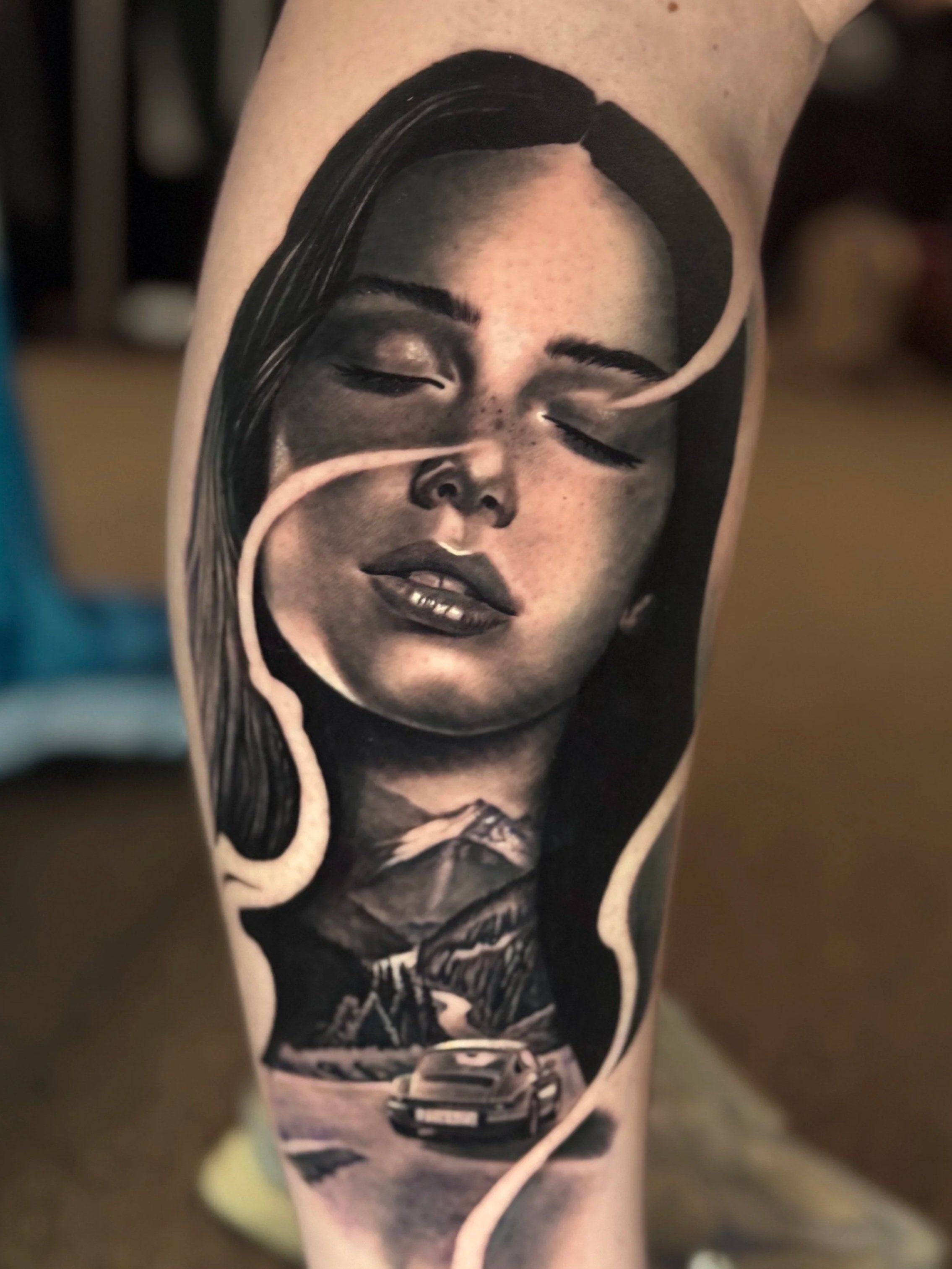 A black-and-white tattoo of a woman with long hair and a gentle smile, wearing a necklace, with a sports car at the bottom of the tattoo.