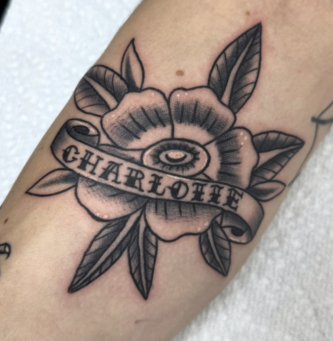 A tattoo of a flower with a banner bearing the name "Charlotte," surrounded by leaves.